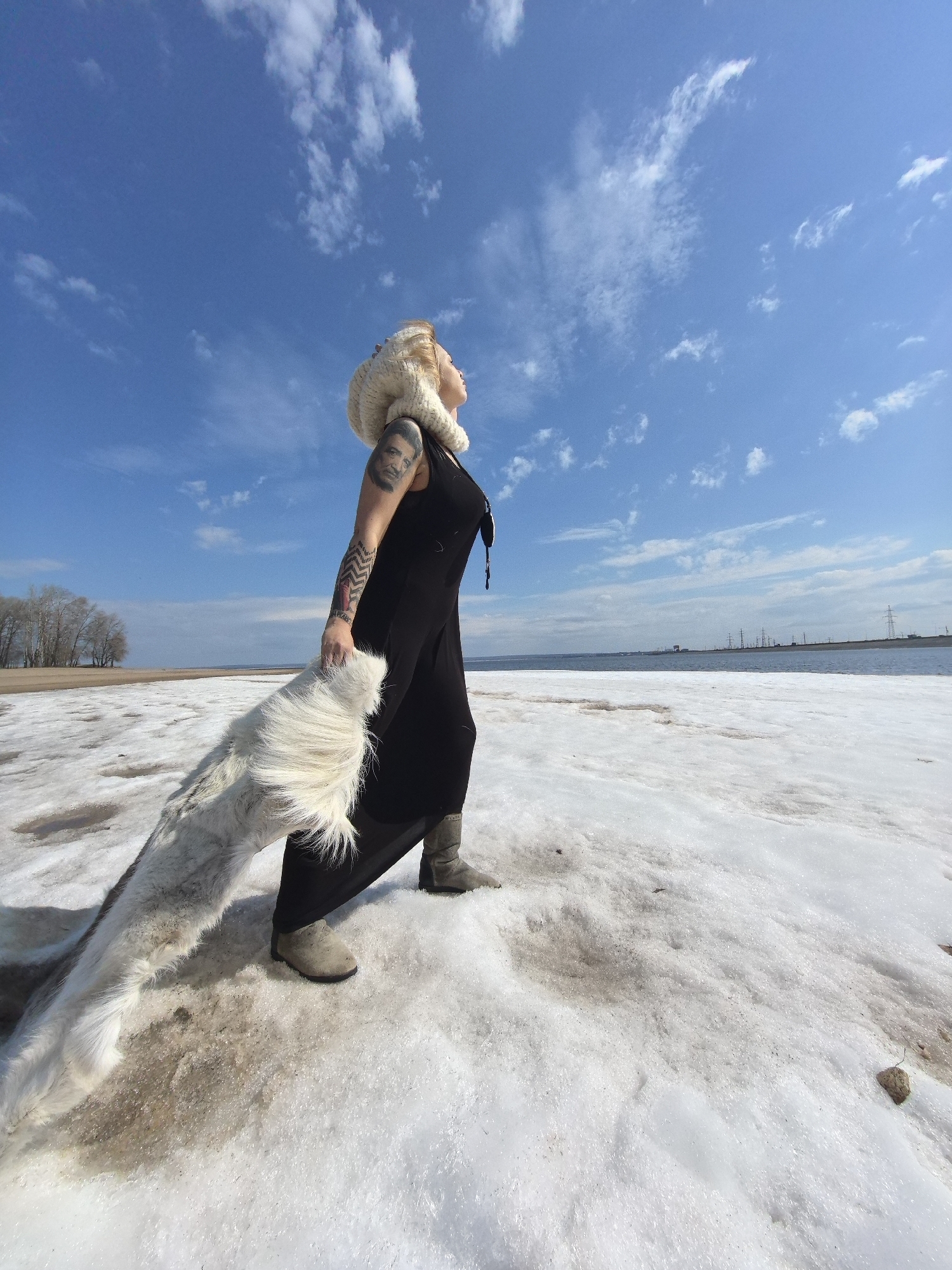 A female figure stands on a thawing landscape of patchy snow and dirt under a bright blue sky with scattered white clouds. She is viewed from a low angle, looking upwards and to her right, with her head slightly tilted back. She has blonde or light-colored hair and wears a large, fluffy white head covering that also appears to function as a scarf. Her attire consists of a long, loose-fitting black dress and grey boots. Distinctive tattoos are visible on her left arm, including a portrait of a man on her bicep and geometric patterns on her forearm. She holds a long, thick white furry item, possibly a pelt or an elaborate scarf, which trails along the ground behind her.
In the background to the left, a line of bare trees suggests a late winter or early spring setting. To the right, a wide body of water, likely a river or lake, stretches into the distance, with several power lines and transmission towers visible on the far bank. The ground beneath her is a mix of melting snow, ice, and exposed brown earth. The bright sunlight indicates it is daytime, with clear weather. The scene evokes a transitional period in a northern climate, consistent with the given location context of Noviy, Russia. No visible text is present.