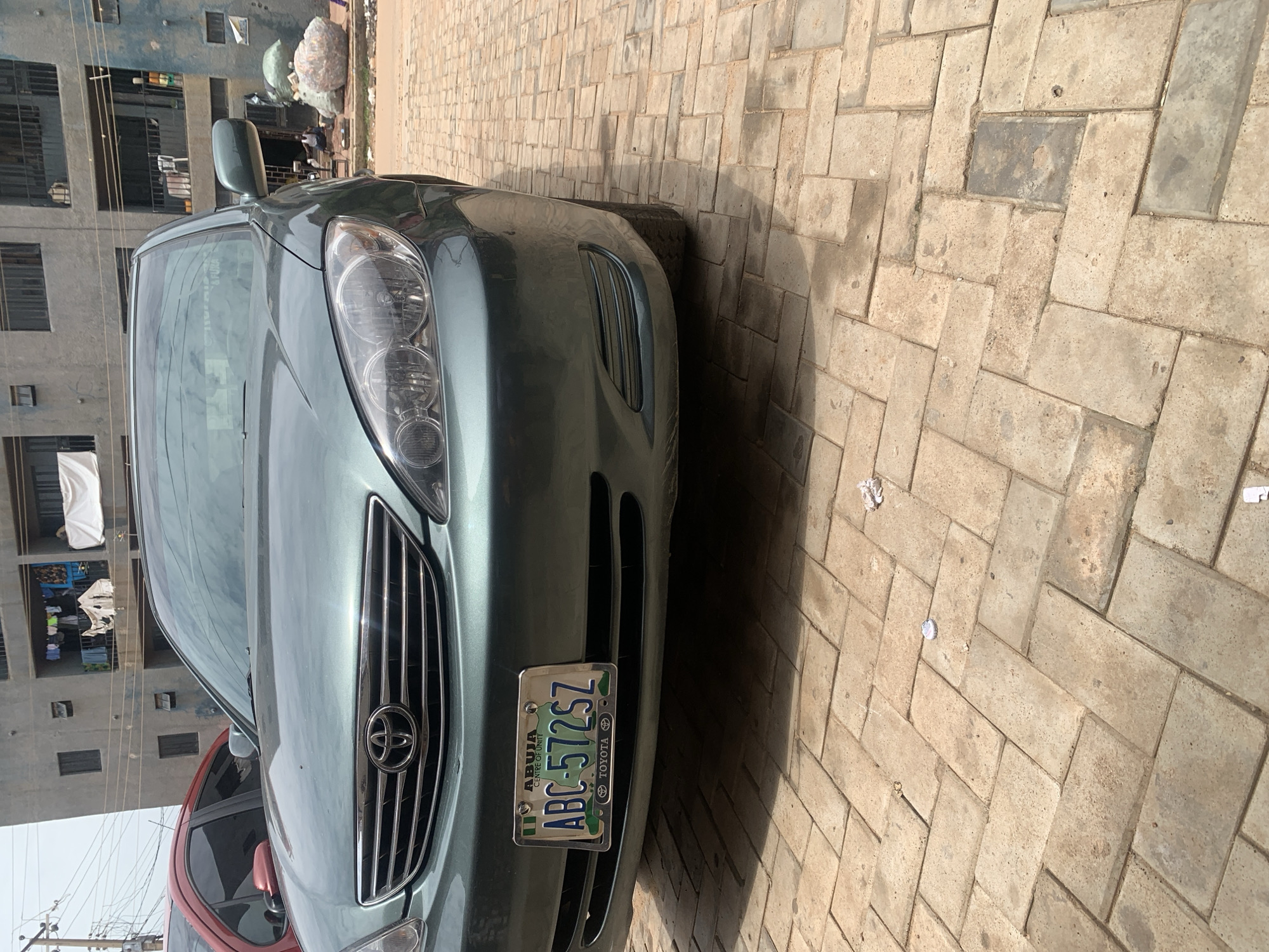 The image shows the front of a dark green Toyota Camry parked on a brick-paved surface. The car's license plate reads "ABC-572SZ" and indicates "ABUJA CENTRE OF UNITY TOYOTA". In the background, a multi-story concrete building is visible with laundry hanging from balconies. To the right of the car, several large piles of compressed materials, possibly recyclable waste, are stacked on the ground. A person is sitting on a low stool near these piles. The overall setting appears to be an urban environment in Nigeria, likely within or near Dadi Riba, given the context. The scene is a static view of parked vehicles and urban infrastructure.