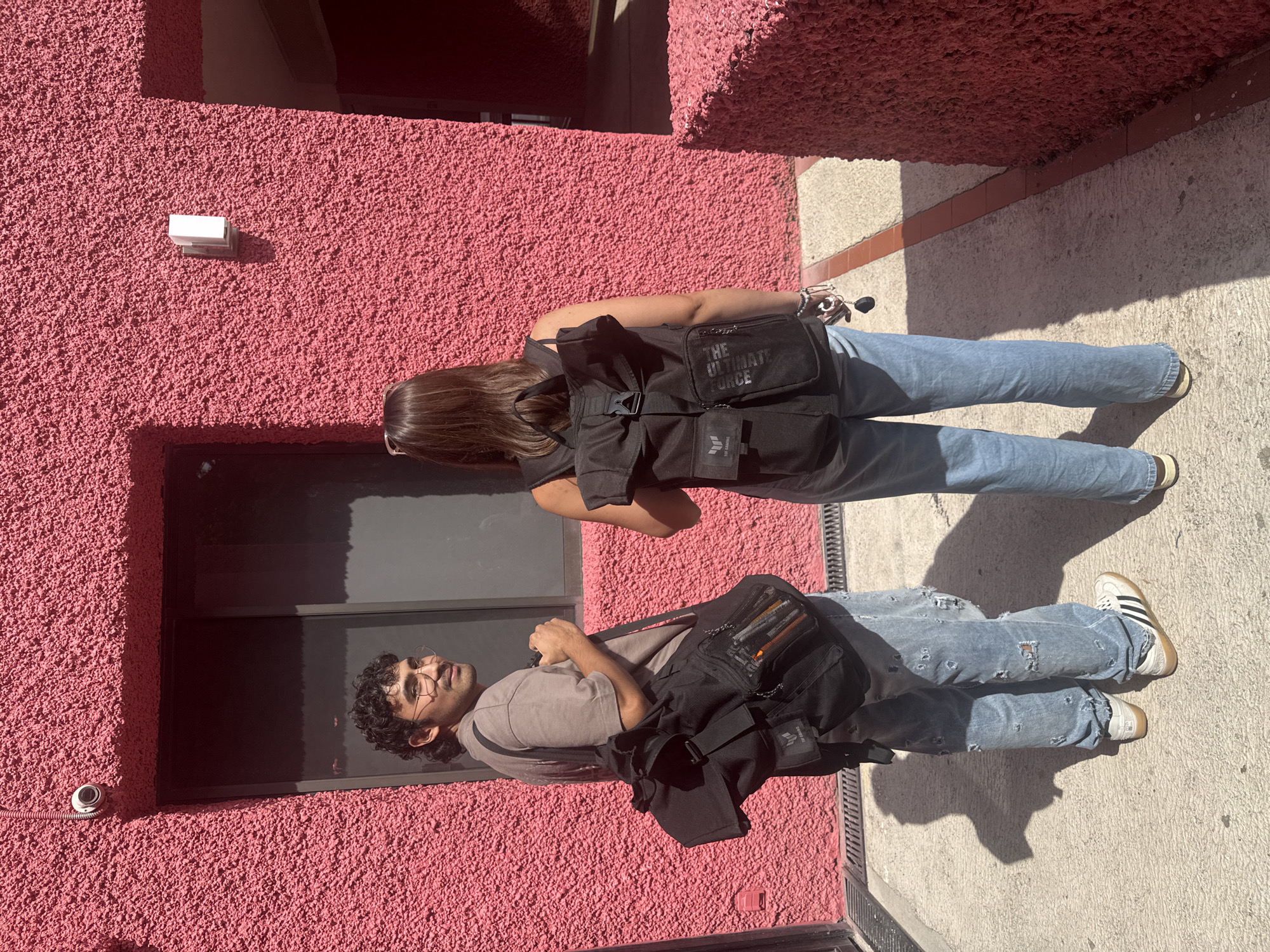 Two individuals are standing on a concrete sidewalk adjacent to a pink textured wall. One person, seen from the back, has long brown hair and is wearing a black backpack with the words "THE ULTIMATE FORCE" printed on it, light blue jeans, and light colored shoes. The other person has dark curly hair, wears glasses, and is wearing a light-colored shirt, light blue distressed jeans, and white sneakers with black stripes. They also have a black messenger-style bag. The location appears to be outdoors, with strong sunlight creating sharp shadows. Based on the architectural style and color palette, the scene is likely located in the city of San Andrés Cholula, Mexico.