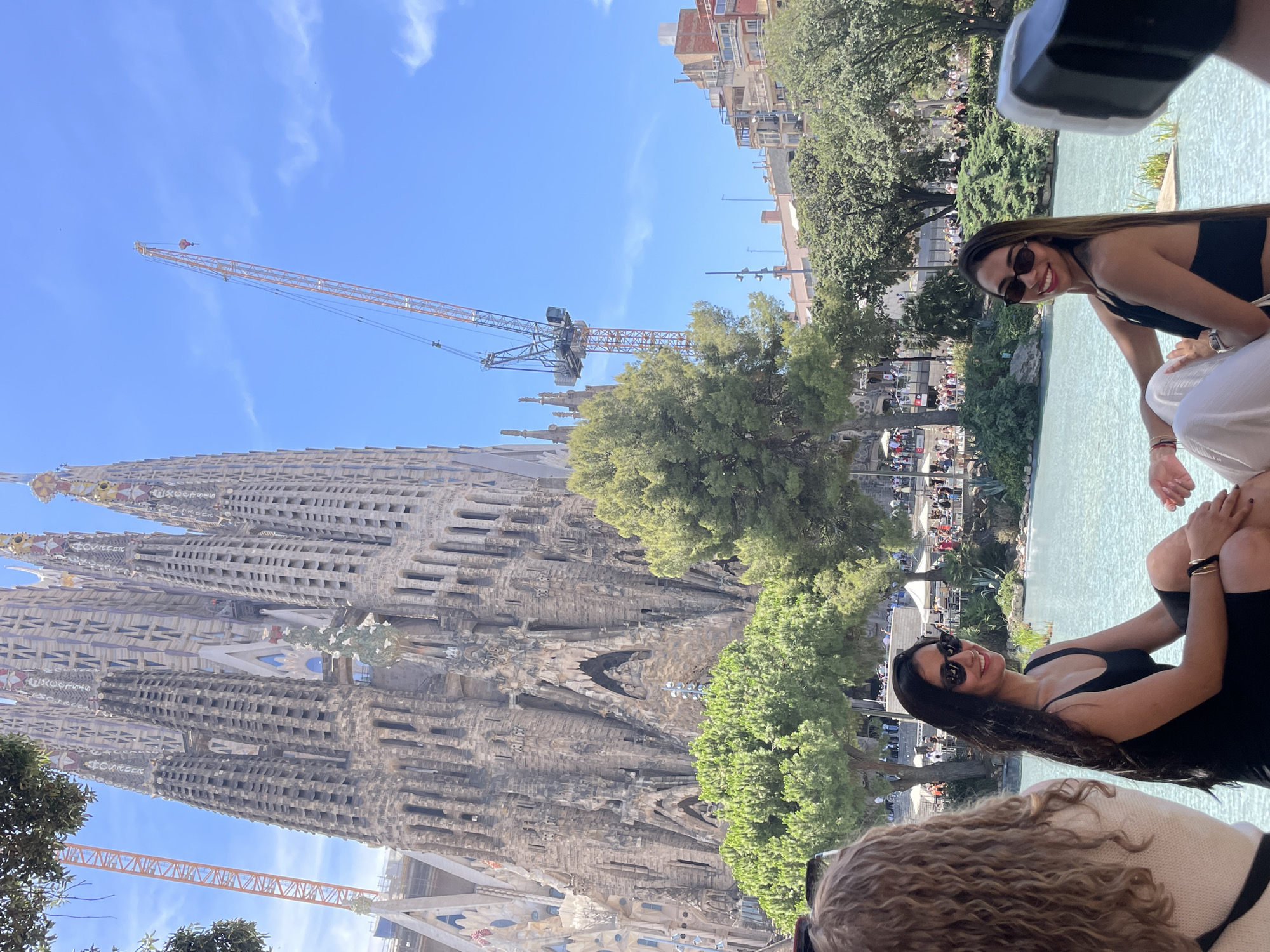 This media file shows a daytime scene in Barcelona, Spain, likely during fair weather given the bright sunlight and clear blue sky. In the background, the iconic Sagrada Familia, a large and intricate basilica, dominates the view. Its unique, ornate architecture, characterized by numerous spires and detailed facades, is clearly visible. Construction cranes are present near the basilica, indicating ongoing work.
In the foreground, two women are seated near a body of water, possibly a fountain or pool. One woman, wearing sunglasses and a black top, is looking at the camera and smiling, suggesting a relaxed and happy mood. The other woman, in the very foreground, has curly hair and is partially visible. The setting appears to be a public area or park, with trees and other people visible in the mid-ground, suggesting it's a tourist attraction or a lively public space. There is no visible text in the image.