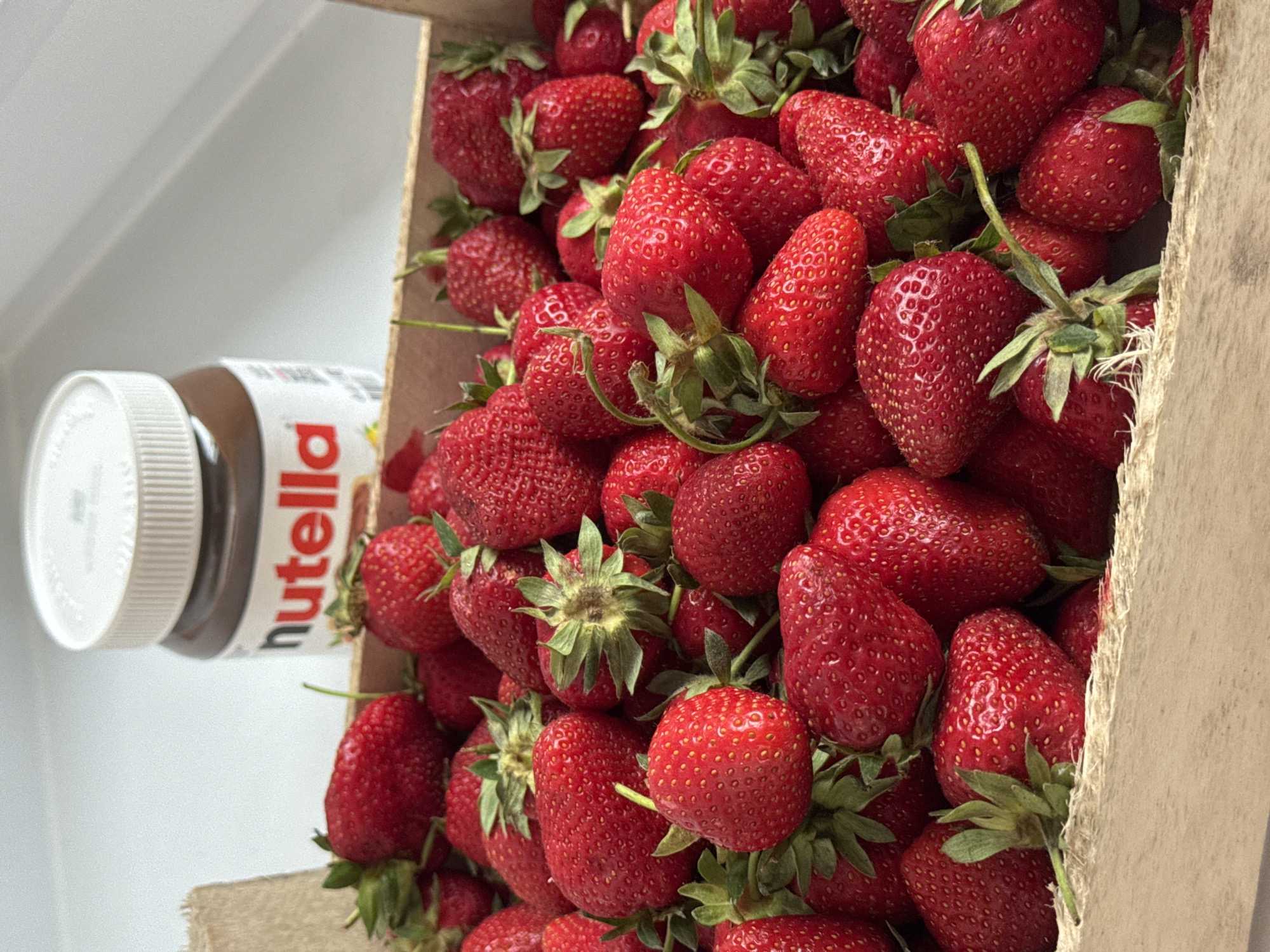 A carton of strawberries sits next to a jar of Nutella. The strawberries are bright red and appear fresh. The Nutella jar is partially visible, with its white lid facing the viewer. The photograph is likely taken indoors, given the lighting and lack of visible outdoor elements. The location, Izhevsk, Russia, cannot be verified from the visual information provided.