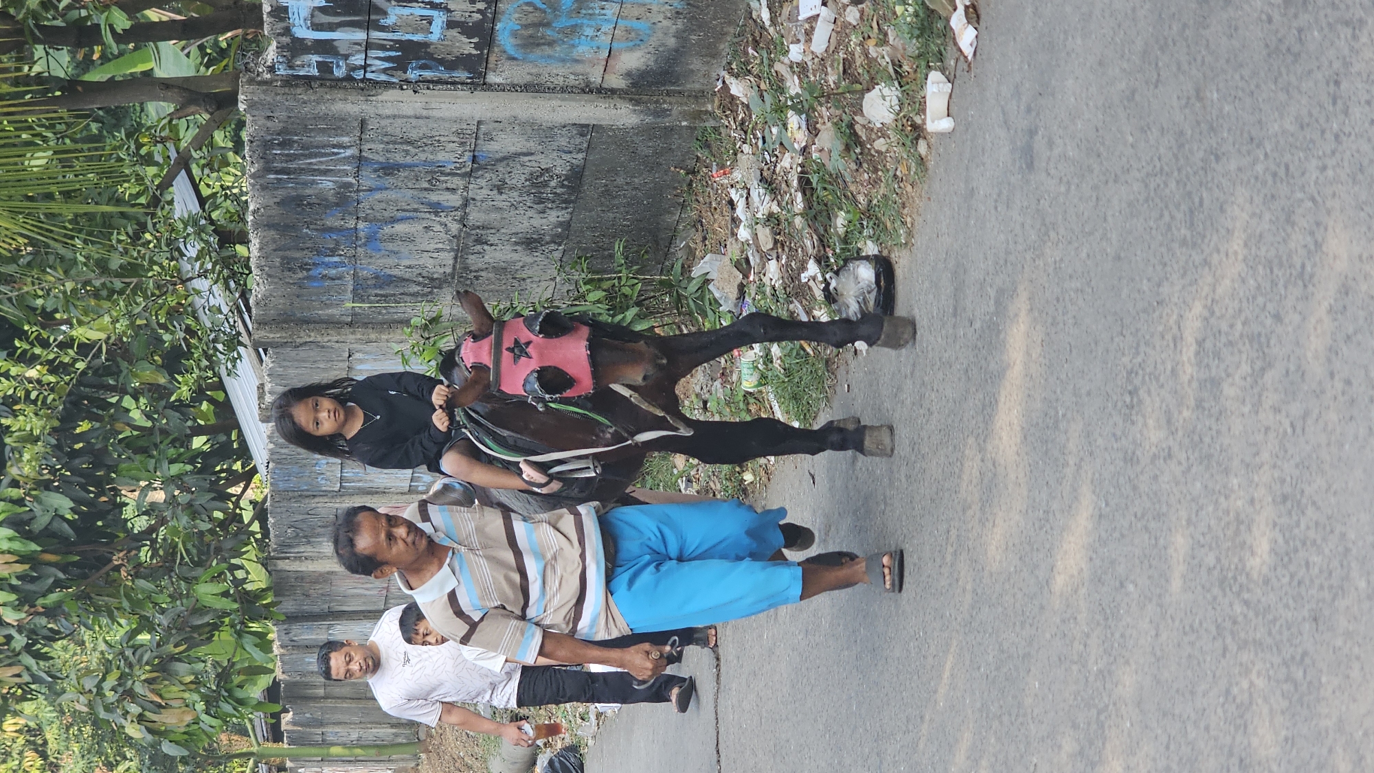 The image shows a street scene in Depok, Indonesia. A young girl wearing a black shirt rides a dark brown horse adorned with a pink mask. An adult man in a striped polo shirt and blue pants walks beside the horse, holding its reins. Another man in a white t-shirt and a child in a white shirt are visible in the background, walking on the left side of the street. The horse is walking on an asphalt road, with a concrete wall and overgrown vegetation on the right side of the street. The wall has graffiti on it. The ground beside the road is littered with trash. The overall scene depicts a casual outdoor activity in an urban environment.