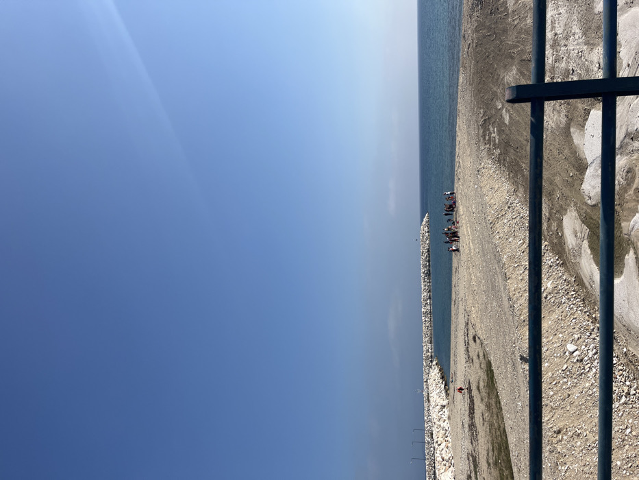 The image shows a beach in Limenaria, Greece. The location is a small bay near the harbor, identified by the coordinates 40.626 latitude and 24.573 longitude. The foreground is a sandy beach with a large concrete structure, or jetty, in the middle. There are white rocks on the structure. The background is a blue ocean. The horizon is marked by a small strip of white sand. The sky is clear and blue. A group of people are gathered on the beach, interacting with each other, near the jetty. There is a large black metal railing in the foreground that is partially obscuring the view of the beach. The sand on the beach is light brown and rough, with some rocks and debris visible. The image appears to have been taken during the day.