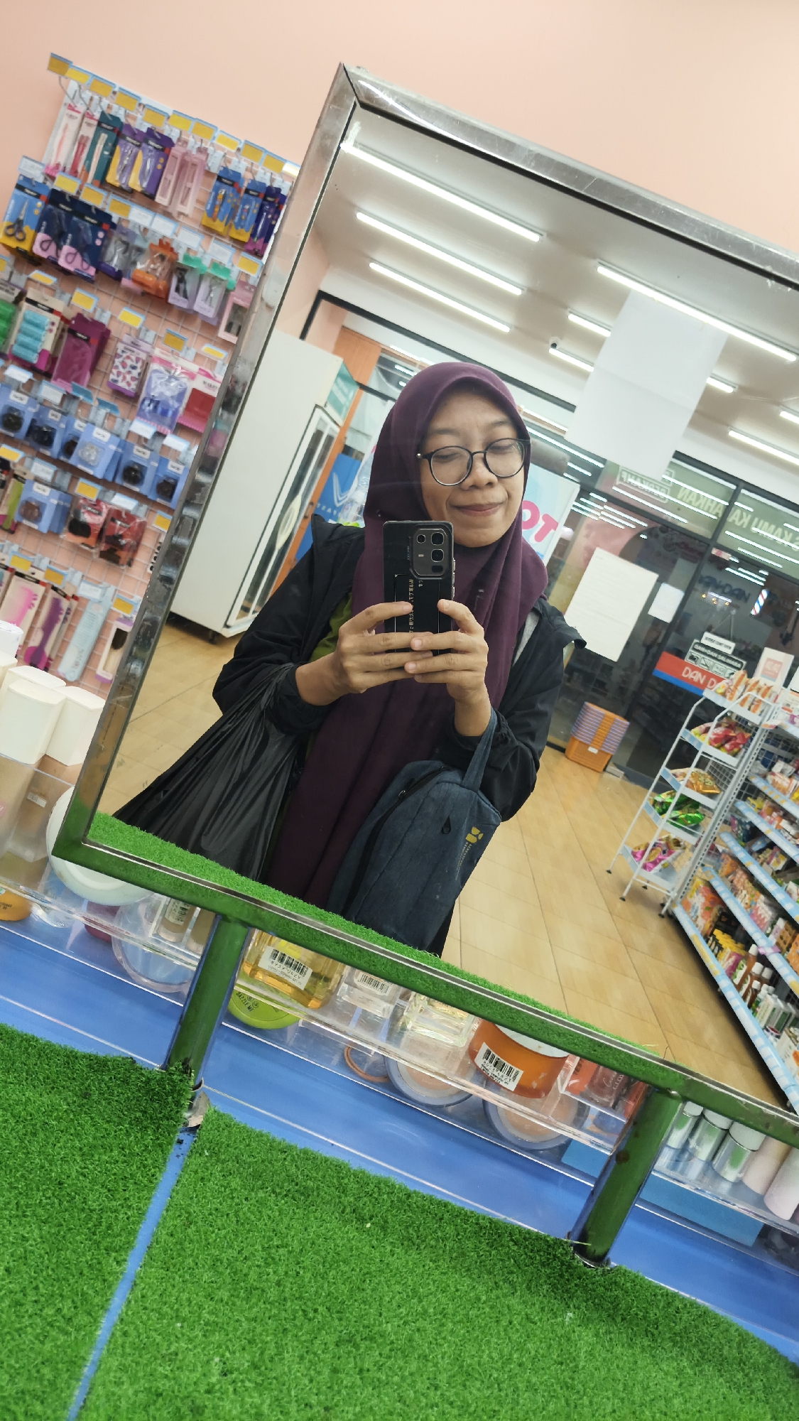 The image is a mirror selfie taken by a woman wearing a purple hijab and glasses in what appears to be a convenience store. She is holding a smartphone to take the photo. She is wearing a black jacket over a green shirt. In her left hand, she holds a black plastic bag, and in her right hand, she holds a dark blue tote bag. Reflected in the mirror, she is standing in an aisle of the store, with shelves of products behind her. The store appears to be well-lit with fluorescent lights. The floor is tiled. The shelves are stocked with various items, including personal care products, snacks, and drinks. The presence of a prominent mirror suggests the photo was taken near the checkout counter or an aisle with a large mirror for security or customer convenience. The overall environment suggests a typical retail setting within a city. Considering the context provided, the location is a convenience store in South Jakarta, Indonesia.