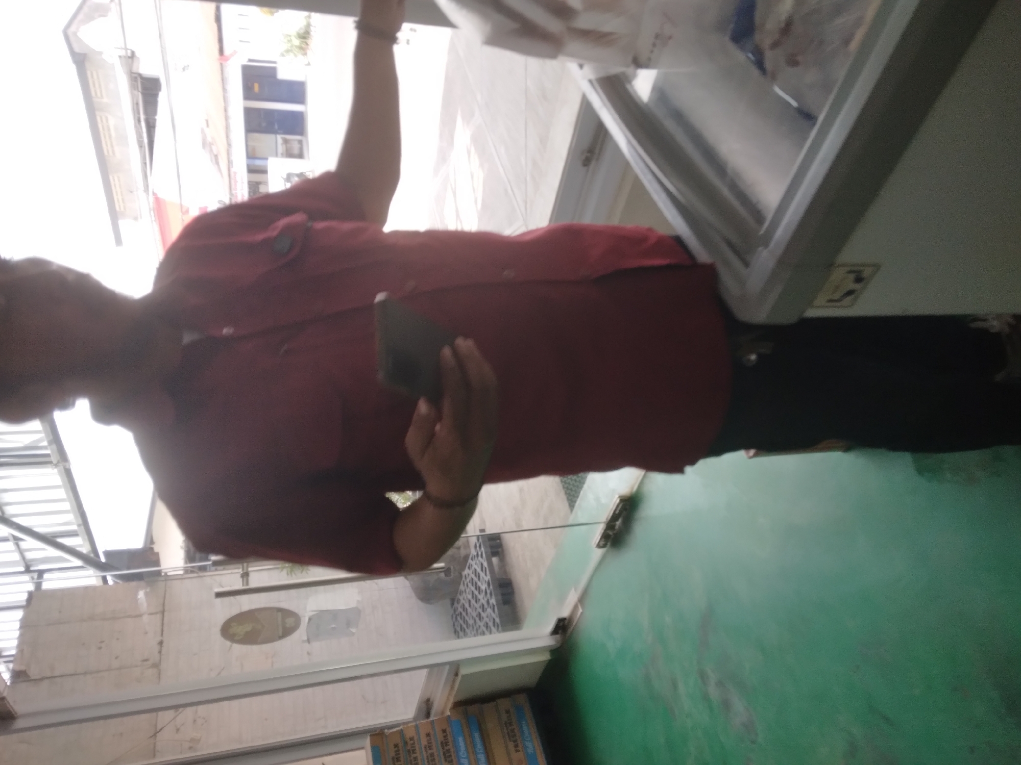In Sokaraja, Indonesia, a person wearing a red collared shirt and black pants stands inside a commercial establishment. They are holding a dark-colored mobile phone in their right hand. To the left of the person, several stacked boxes labeled "FRESH MILK" are visible, suggesting a dairy or grocery store setting. In the foreground, a chest freezer with a glass lid contains frozen goods. The floor is a reflective green color. Beyond the person, a glass door and a glimpse of an outdoor area with buildings can be seen.