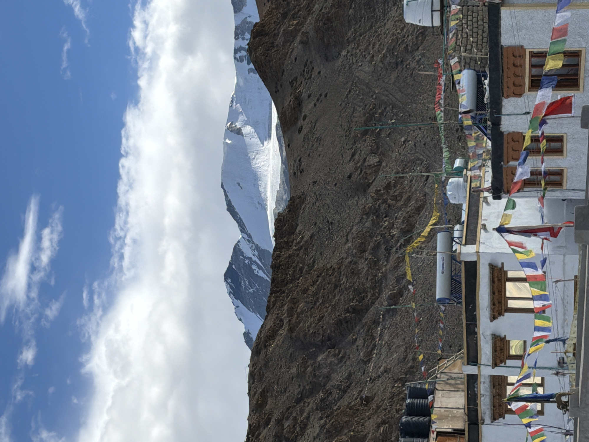 In Karzok, India, a village situated in the high-altitude region of Ladakh, a cluster of buildings with white walls and wooden window frames is seen against a backdrop of rugged, barren mountains. Prayer flags in vibrant colors of blue, white, red, green, and yellow are strung across the rooftops and between the buildings, fluttering in the breeze. Several water tanks, likely for domestic use or solar heating, are visible on the roofs, alongside solar water heaters. The mountains behind are stark and rocky, with snow-capped peaks dominating the horizon under a partially cloudy blue sky. The overall scene depicts a peaceful, remote settlement in a dramatic mountainous landscape.