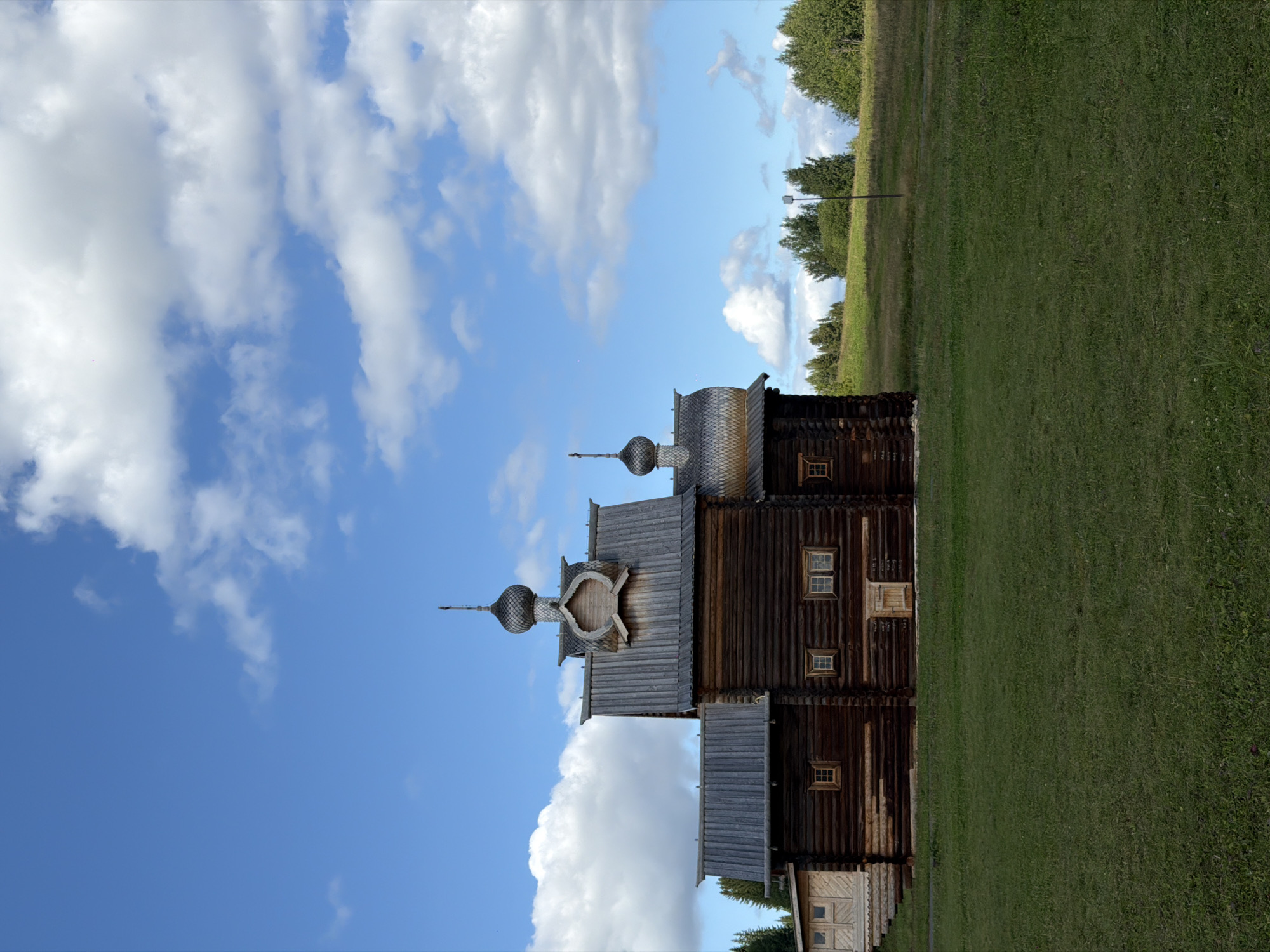 An old, dark wooden church with multiple roofs and a domed steeple stands on a grassy field under a partly cloudy blue sky. The church has several windows and a wooden door. To the left of the main structure is a lighter colored wooden annex with windows. The background consists of a gentle, rolling green hill with scattered trees, and a line of darker green trees in the distance. The scene appears to be located in a rural or historical park setting.