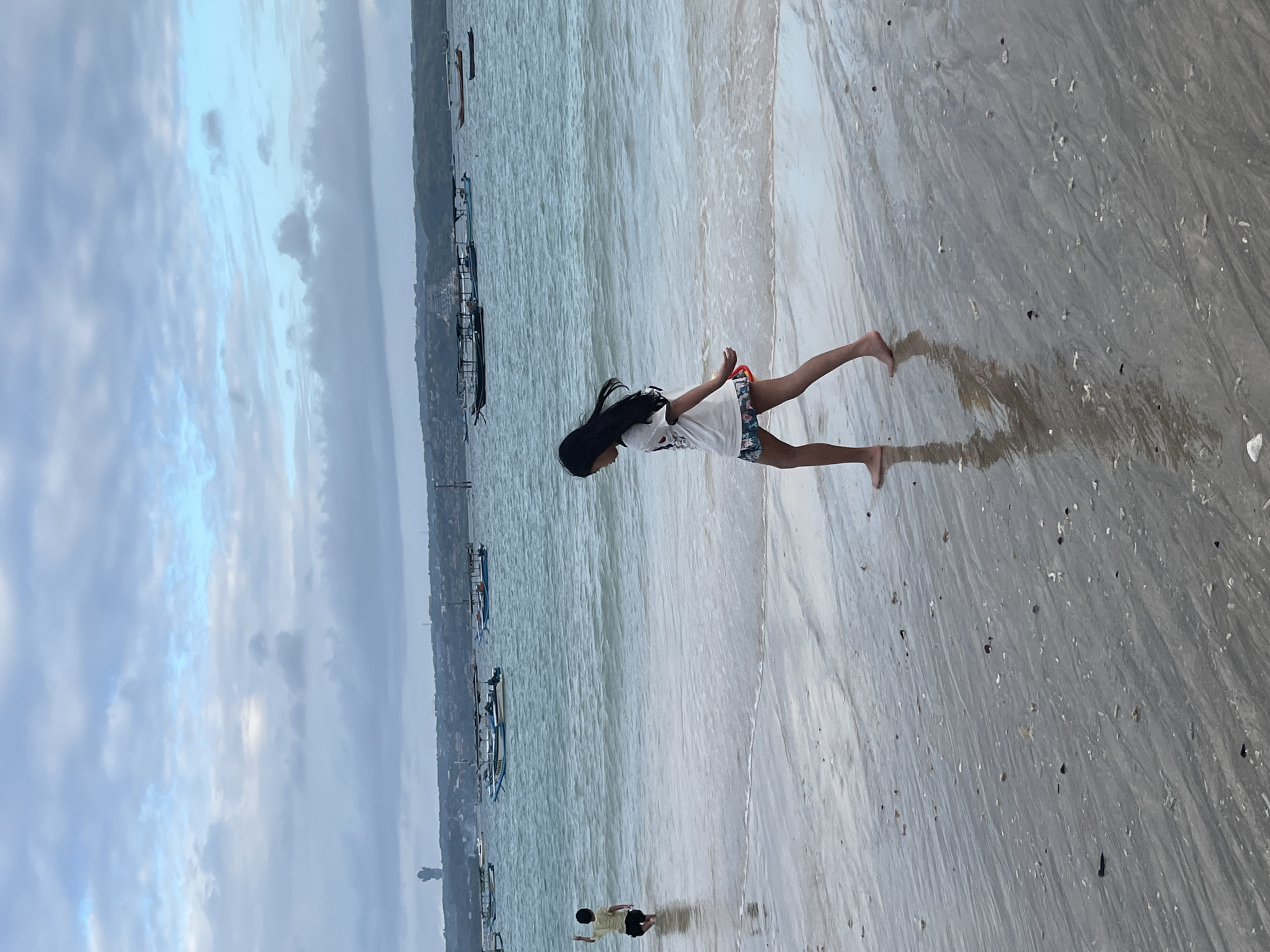 A young girl with long dark hair, wearing a white t-shirt and patterned shorts, walks along a sandy beach in Kuta, Indonesia. Her reflection is visible in the wet sand. In the background, a child in a yellow shirt wades in the ocean, facing away from the camera. Several traditional Indonesian outrigger boats are anchored on the turquoise water, with a distant, hazy coastline visible. The sky is cloudy with patches of blue. The tide is low, revealing a wet sandbar littered with shells.