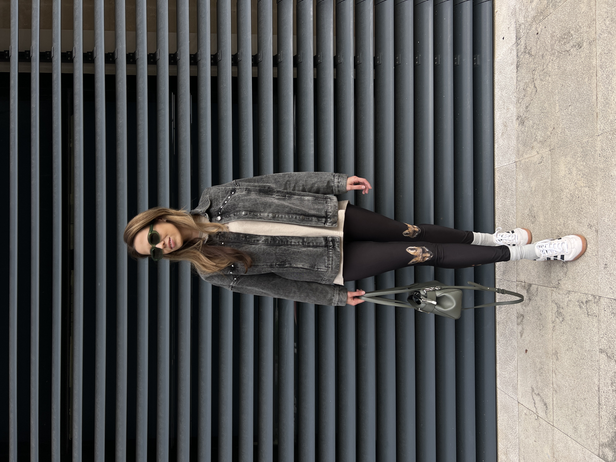 A woman is posing against a facade consisting of closely spaced vertical gray bars. She is wearing a faded gray denim jacket, a white top, black leggings with an animal print on the thighs, white socks, and white sneakers. She also sports sunglasses and holds a small green purse. The setting is a city street in Bar, Montenegro, as evidenced by the local architectural style of the facade and the stone paving on the ground adjacent to the wall.