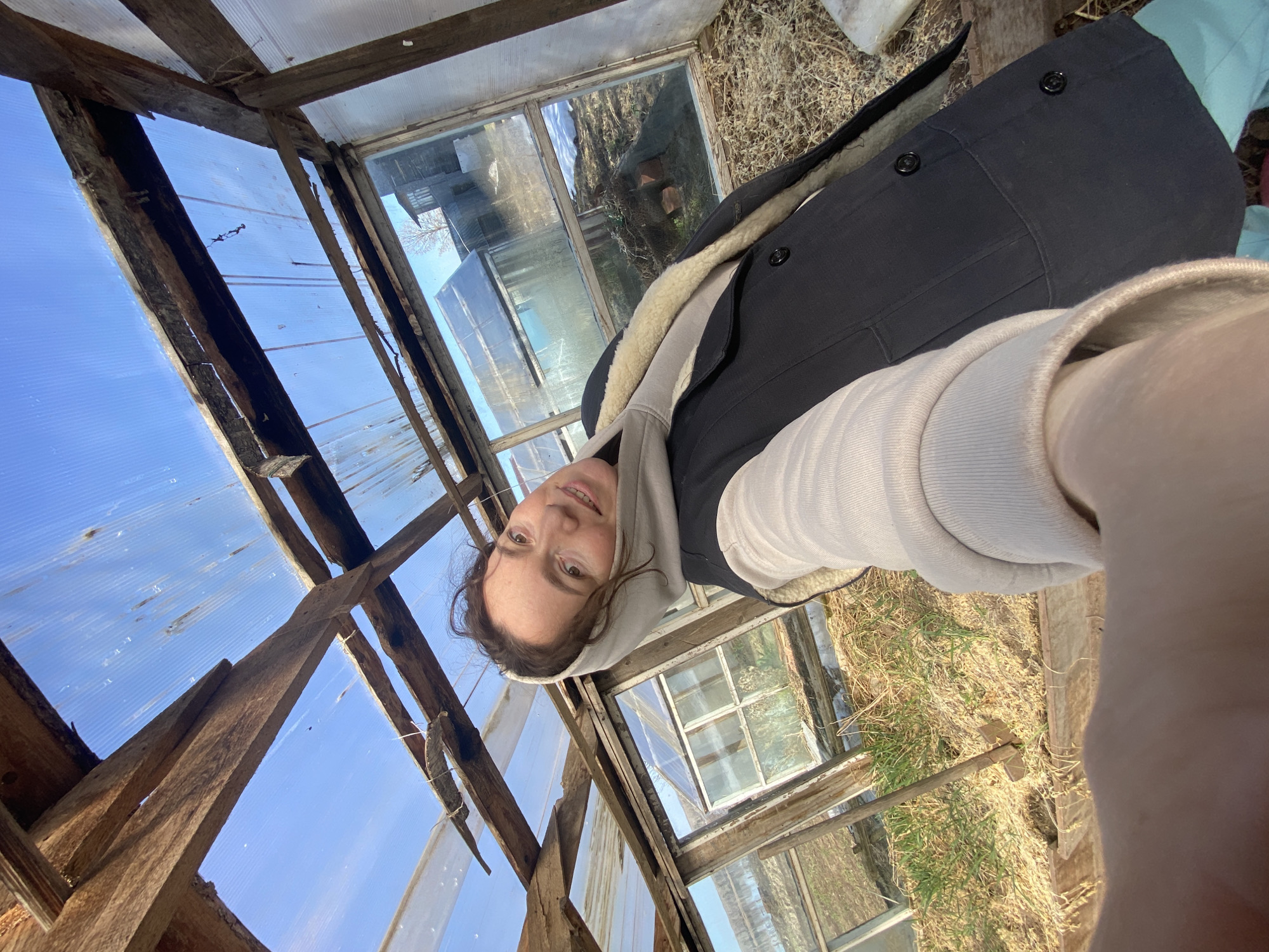 The image shows a woman in a grey hooded sweatshirt and a dark coat with a light-colored fur lining, apparently taking a selfie inside a dilapidated greenhouse. The structure is made of weathered wood and transparent material, possibly plastic or glass, that forms the walls and roof. Sections of the greenhouse show the exterior, revealing an environment with dry grass and other structures. The sky is visible through gaps in the greenhouse structure. The photo seems to be taken at an angle, with the person holding the camera at arm's length. The location is Ольховка, Russia.