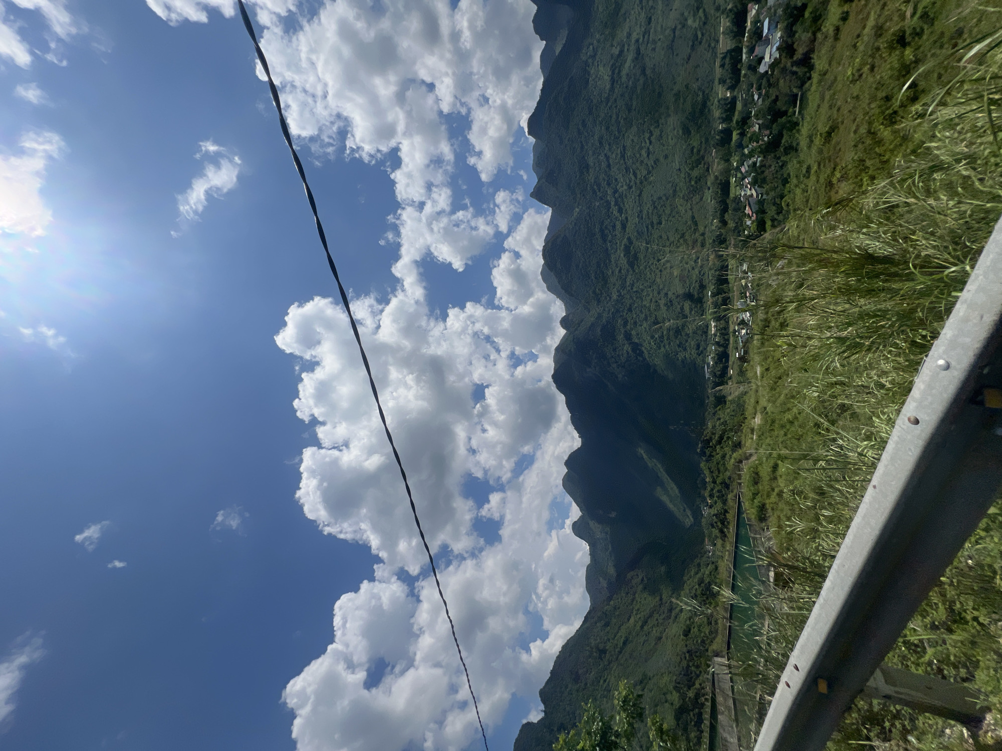 The image depicts a scenic landscape in Lùng Tám Commune, Vietnam, under a bright blue sky dotted with fluffy white clouds. In the foreground, a metal guardrail runs diagonally across the frame, weathered and showing signs of rust. Beyond the guardrail, tall, wild grasses sway gently, giving way to a body of water with a greenish tint, possibly a reservoir or pond, bordered by concrete structures. Further in the background, a lush green valley unfolds, leading to a range of densely vegetated, jagged mountains that dominate the horizon. Nestled within the valley, a small village with houses featuring red roofs can be seen amongst the trees. A dark, thin wire stretches diagonally across the top portion of the image, obscuring a small section of the sky and the tops of the mountains. The sun is shining brightly, casting a slight glare in the upper right quadrant of the sky. No people or animals are visible in the scene. The overall impression is one of natural beauty and a tranquil rural setting.