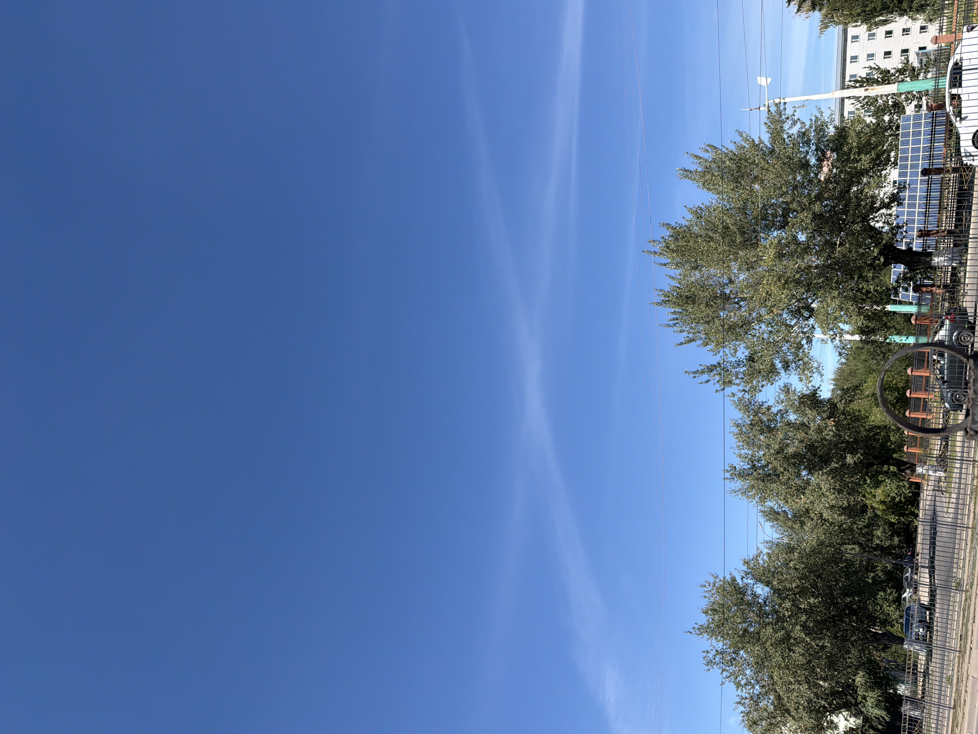 The image shows a clear blue sky with thin, wispy clouds. In the lower portion of the frame, there are trees, a street, parked cars, a fence, and buildings. The environment appears to be urban. Based on the context provided, this scene is located in the city of Pavlodar, Kazakhstan.