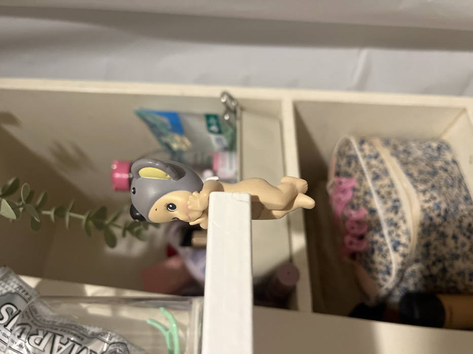 A small, nude-colored plastic figurine of a baby dressed as a gray koala lies atop a white divider in a shelf. The shelf compartments hold various items including dental floss, cosmetics, a floral patterned bag, and toiletries. A green leafy stem is to the left of the figurine. The background is a soft white, potentially the inside of a cabinet.