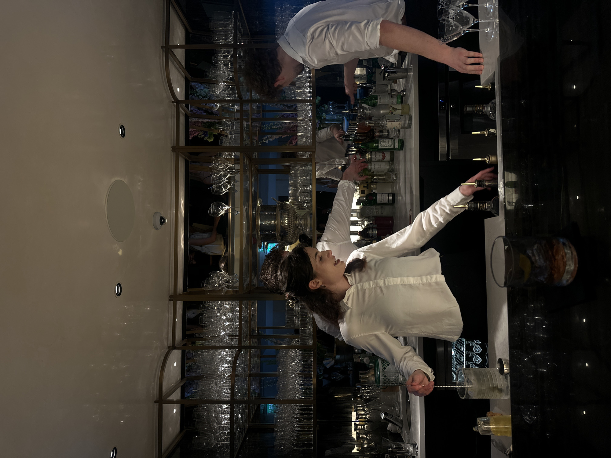 Here is a description of the image:
**Scene**: The image appears to be taken inside a high-end cocktail bar in London, United Kingdom. The scene is dimly lit with a focus on the illuminated bar area.
**People**: Two individuals are present. One is a young woman with dark, wavy hair wearing a white button-down shirt. She is actively working behind the bar, stirring a drink in a glass with a bar spoon. The second person, also wearing a white shirt, is partially visible at the top of the frame and appears to be another bartender.
**Objects**: A well-stocked bar is visible, featuring multiple shelves of liquor bottles and glassware. Specific items include various types of cocktail glasses (martini glasses, coupes, etc.), liquor bottles of different shapes and sizes, bar tools (bar spoon, mixing glasses).
**Actions/Interactions**: The primary action is the female bartender preparing a cocktail. She is focused on stirring a drink, suggesting precision and expertise.
**Environment**: The bar area features a polished black countertop with a reflective surface. A decorative gold-toned metal shelving unit displaying the glassware adds to the upscale ambiance. The general environment indicates a sophisticated cocktail bar.