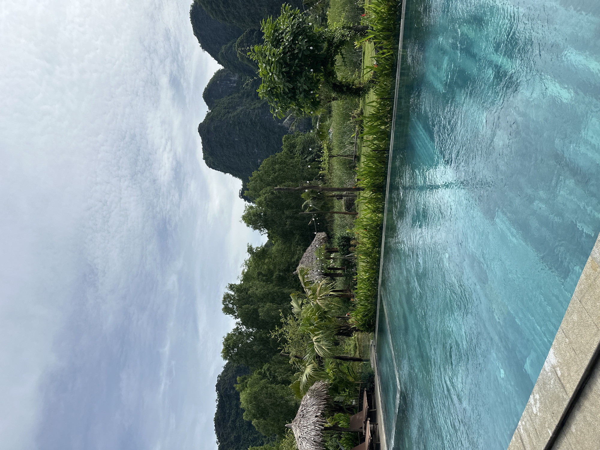 This image shows a swimming pool in the foreground, with lush greenery and karst mountains in the background. The swimming pool is rectangular and appears to be an infinity pool, with the water flowing over the edge. The water is a clear turquoise color, and there are some lounge chairs visible on the deck next to the pool. In the background, there are two thatched-roof huts, palm trees, and other tropical vegetation. Beyond the vegetation, there are several large, jagged karst mountains covered in trees. The sky is cloudy with some patches of blue. The scene suggests a resort or a private residence in a tropical or subtropical location. The caption indicates this is in Nam Hoa Lư Ward, Vietnam, a region known for its natural beauty and limestone karsts.