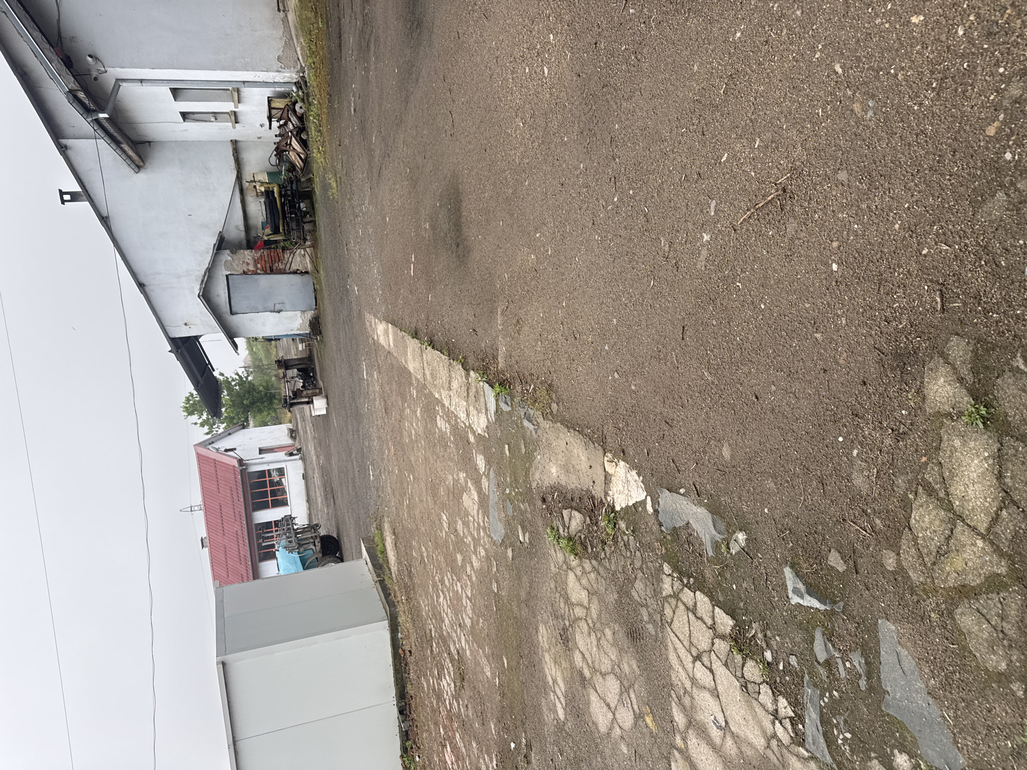 This media file captures an outdoor scene, likely an industrial or agricultural yard, in Sarnevo, Bulgaria. The image depicts a ground-level view looking towards a cluster of buildings under an overcast sky.
The foreground features a mixed ground surface. On the right, it is a dark, coarse asphalt or compacted dirt path. This transitions to the left into an area of broken and cracked concrete or paving stones, with small patches of grass and weeds growing through the fissures. A distinct concrete strip separates these two ground types.
Several buildings are visible. On the far left, a large, light-colored (white or light grey) building with a sloped roof and a dark gutter system dominates part of the scene. A security camera is mounted on its upper left corner. Below this building, a grey metal door is present, and a pile of miscellaneous items, possibly scrap metal or discarded machinery, is accumulated nearby. Further back and slightly to the right, another white building with a distinctive red tiled roof and large windows is visible. In front of this building, outdoor equipment, including a blue tarp covering an object and parts of a small vehicle or machine, can be seen. Towards the left foreground, a large, modern-looking grey rectangular structure, possibly a container or part of a larger building, is partially visible.
The sky is uniformly grey and overcast, indicating a cloudy day. There are no harsh shadows, suggesting either early morning, late afternoon, or a heavily clouded mid-day. Power lines are visible stretching across the mid-ground against the sky. No people are present, and no specific activity is taking place. There is no visible text in the image.