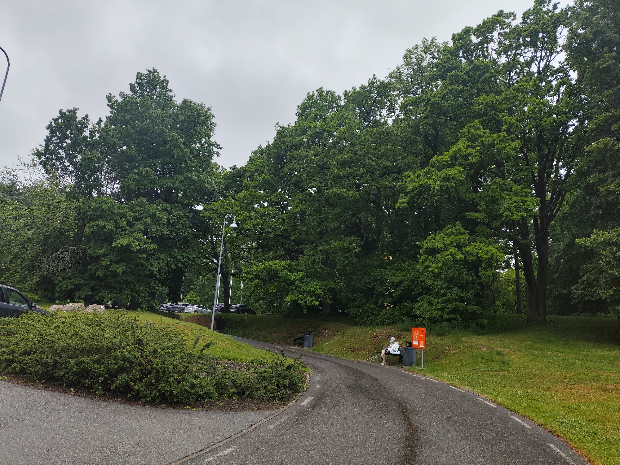 The image shows a paved walking path curving through a park-like setting. Dense green trees line the background, with a clear sky overhead. On the right side of the path, a person is seated on a dark bench, facing away from the camera. The person is wearing a light-colored hooded jacket and dark pants, and is holding a white object in their hands. Adjacent to the bench is an orange mailbox. In the foreground, a manicured hedge borders the path. A parking lot with several cars is visible on the left side of the image, set back from the path and slightly uphill. A street lamp is also present on the left. The scene appears to be within the city of Tallinn, Estonia, judging by the presence of the mailbox which resembles those used in Estonia.