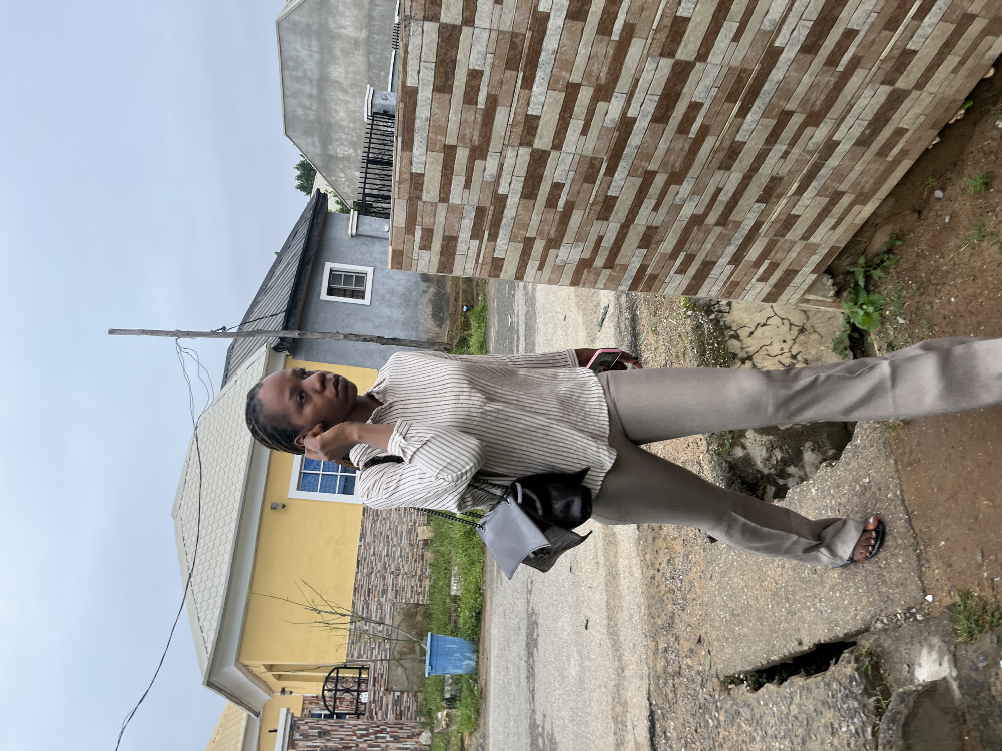 A medium-shot, eye-level, outdoor, full-body photograph shows a young Black woman walking on a cracked concrete pathway. She is wearing a light-colored, vertical-striped button-down shirt and taupe-colored, flared pants. The woman has her hair in braids and is holding her right ear with her right hand. She carries a black and gray shoulder bag over her left shoulder. She is walking towards the right of the frame, with her left foot on the pathway and her right foot in the air. A phone with a pink case is visible in her right hand.
The environment is a residential area, likely in Abuja, Nigeria, with several houses visible in the background. To the left of the woman is a yellow house with a white roof and a blue plastic bin. Further left, there is a dark metal gate. Behind the woman, there are gray houses with dark roofs and windows. A wall with a patterned tile facade, featuring rectangular tiles in shades of brown and beige, runs along the right side of the frame, partially obscuring the background. The ground is dirt and gravel, with some grass and vegetation growing in patches. The sky is overcast.