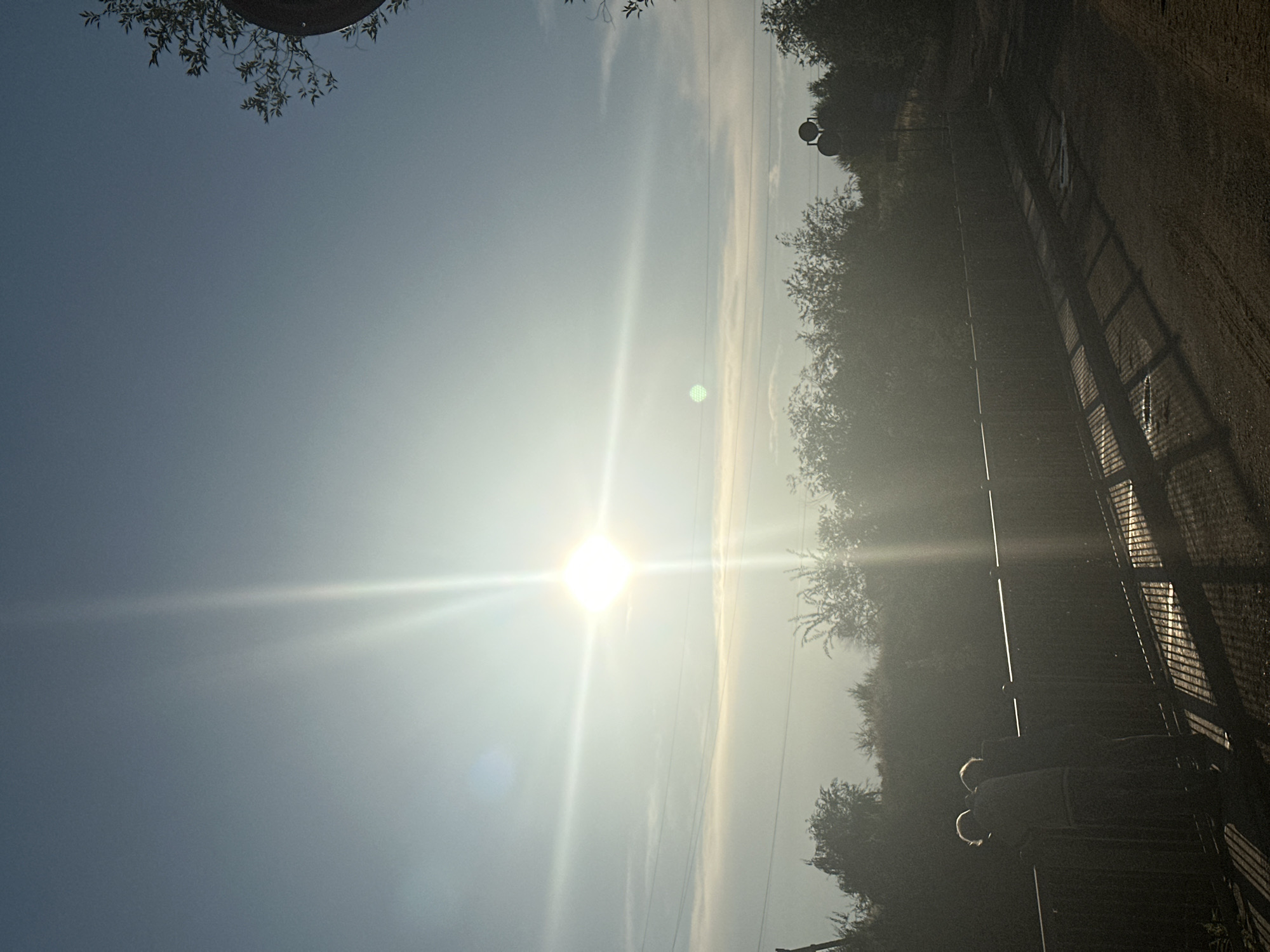 The image depicts a hazy outdoor scene, likely in the city of Energetik, Russia, as indicated by the location tag. The sun is prominent in the sky, creating a bright glare that obscures details in its vicinity. Silhouetted against the bright sky and hazy atmosphere are two figures standing on a bridge or elevated walkway, facing away from the camera. They appear to be leaning on a railing. The foreground shows a dirt or gravel path, casting long shadows. Dense foliage, possibly trees and bushes, lines the background, partially obscuring the horizon. A faint hint of power lines can be seen stretching across the sky. In the upper right portion of the image, a dark, circular object, possibly a sign or part of a structure, is visible amongst the foliage. The overall impression is of a slightly overcast or foggy day with strong sunlight.