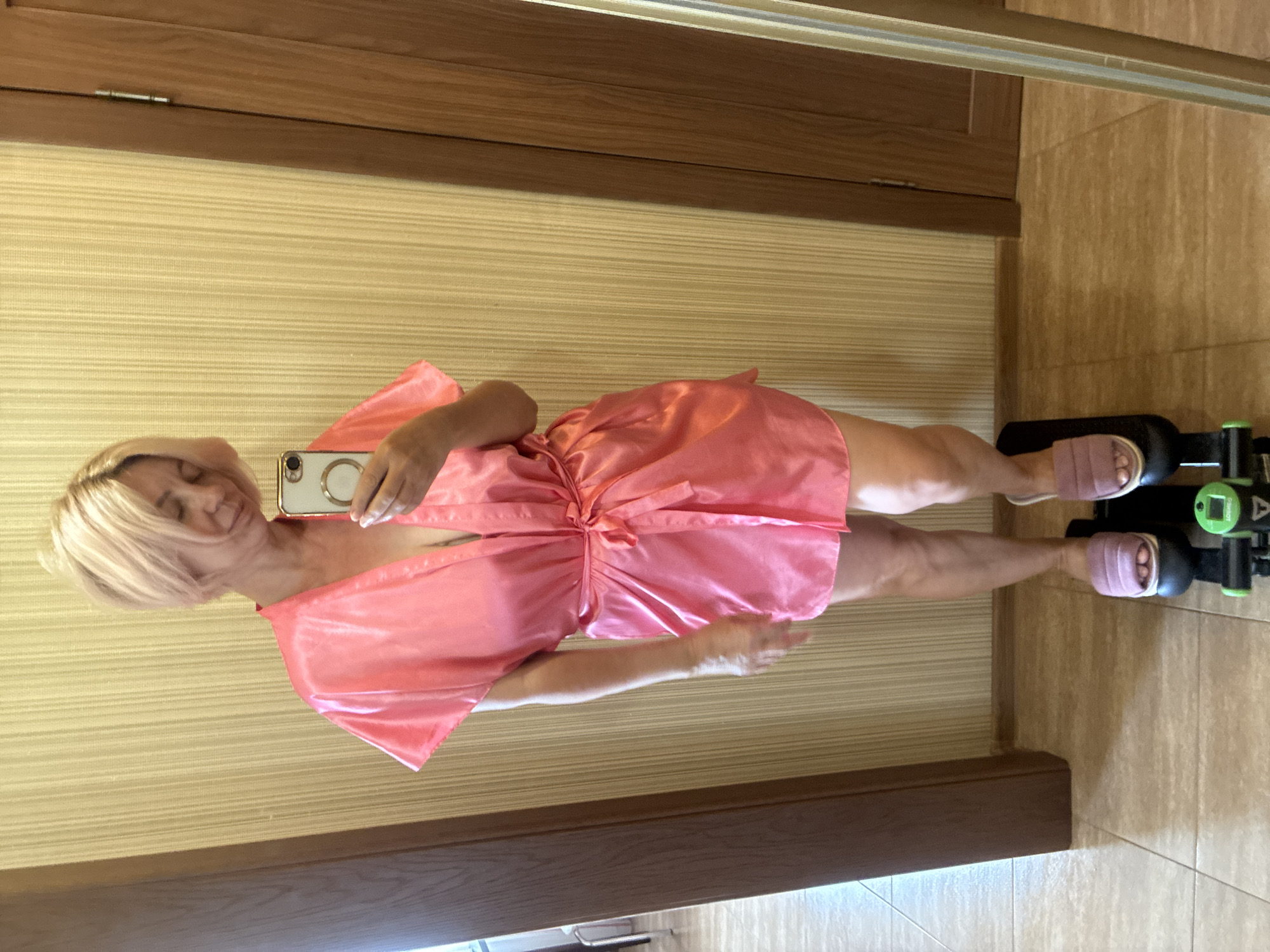 A woman with short blonde hair, wearing a pink satin robe and pink slippers, stands on a small exercise stepper. She is holding a smartphone in her right hand, taking a selfie in a mirror. The background consists of a textured yellow wall and a wooden door. The floor is tiled. The image appears to be taken indoors, possibly in a home or hotel room. There is no indication of the city of Sochi, Russia in the image.