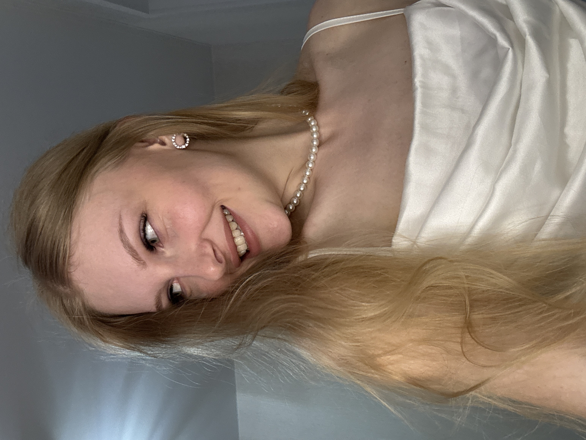 The image is a close-up, waist-up portrait of a young woman with long, blonde hair. She is wearing a white, satin slip dress and a pearl necklace. She is also wearing small, circular earrings. Her head is tilted to the left, and she is smiling, looking away from the camera. The background is a plain, light gray wall. There is no discernible indication of location within the city of Perm, Russia.