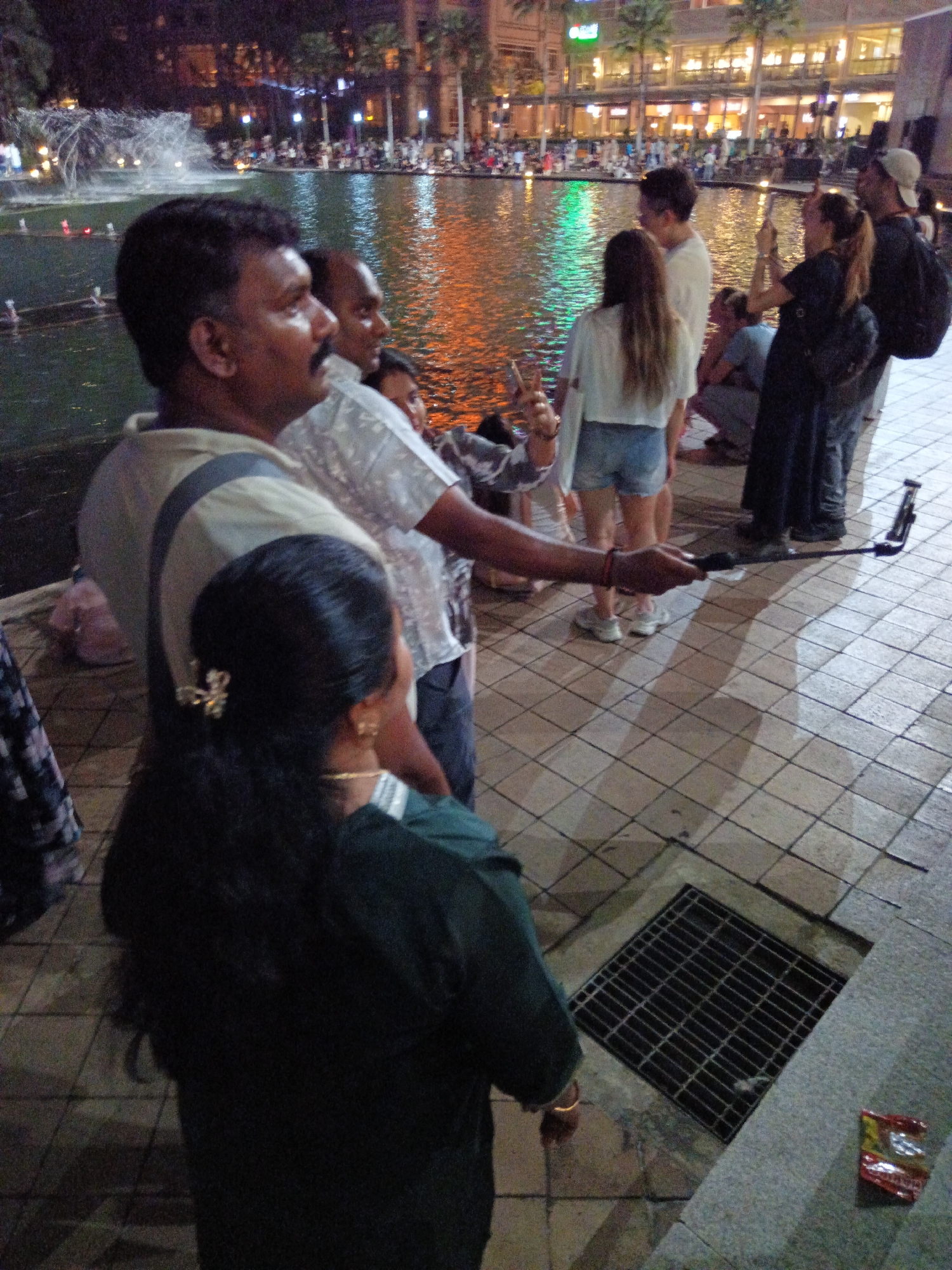 The image depicts a dimly lit outdoor scene, likely in Kuala Lumpur, Malaysia, given the context of the provided crops, at night. In the foreground, a man with dark skin and a mustache is partially visible, facing left, wearing a light-colored shirt with a dark stripe across the shoulders. Next to him, a woman with long, dark hair adorned with a gold hair clip is seen from behind, wearing a dark green outfit. Behind them, other individuals are present, including a man in a white shirt and a person holding a selfie stick with a phone attached. In the background, a body of water with a fountain is visible, illuminated by colorful lights. Beyond the water, there is a building complex with multiple illuminated floors, possibly a shopping mall or entertainment venue, and palm trees. People are scattered around the area, some appearing to be watching the fountain. A metal grate and a crumpled snack wrapper are visible on the tiled ground in the lower right portion of the image. The overall atmosphere suggests a public gathering or event taking place in a city center.