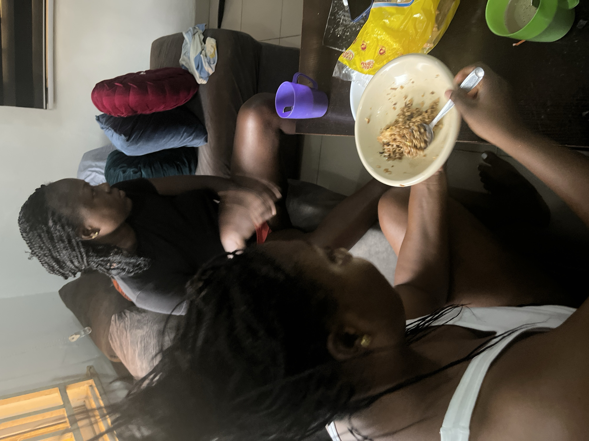 The image depicts an indoor scene, possibly a living room in Abuja, Nigeria, where two individuals are present. In the foreground, a person wearing a white tank top is seated on the floor, holding a bowl of rice with a spoon. Their focus is on the food. In the background, another person with braided hair is seated on a sofa, looking away from the camera. The sofa is adorned with several cushions of varying colors. A coffee table in front of the sofa holds a purple mug, a plastic bag, and a green cup containing a beverage. The environment appears casual and domestic.