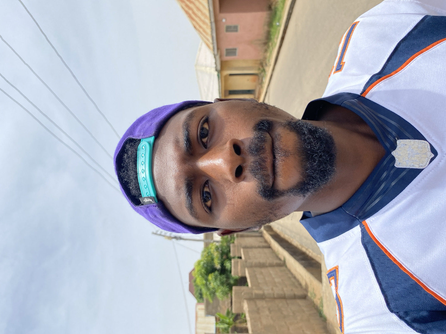A close-up, portrait-oriented selfie shows a man of dark complexion looking directly at the camera. He has a short dark beard and mustache. He is wearing a white sports jersey with navy blue and orange accents; a silver-colored badge is affixed to the collar area. A purple baseball cap is worn backwards, featuring a teal plastic snapback closure. The word "STUSSY" is visible on a tag on the cap, and the number "1" is faintly discernible on the jersey.
The man is positioned outdoors during what appears to be daytime, likely under an overcast sky, as suggested by the bright, diffused light. In the background, thin power lines stretch across the sky. Distant buildings, possibly residential with reddish-brown roofs, are visible, along with some green trees and foliage. A light-colored ground surface, resembling a dirt or unpaved road, runs through the scene, flanked by low concrete or stone structures. The overall setting suggests a casual, outdoor environment, consistent with Jos, Nigeria.