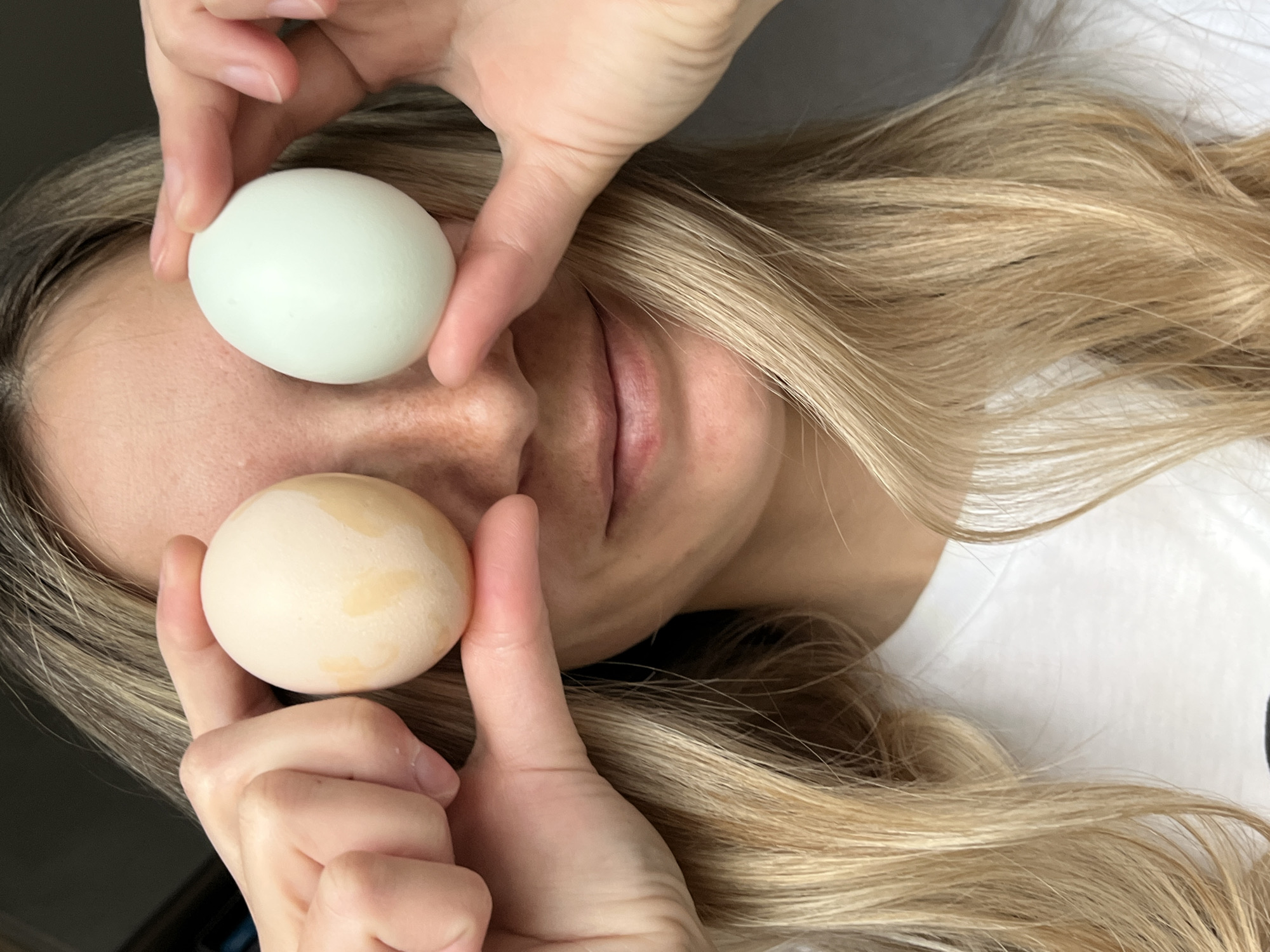 A close-up, vertical photograph depicts a person playfully posing indoors, holding two eggs over their eyes. The individual, likely female, has long, wavy blonde hair that cascades around their head and shoulders. One hand, with fingers visible, holds a smooth, light green-blue egg over the upper part of the face, obscuring one eye and the forehead. The other hand holds a light beige egg, marked with subtle darker spots, over the lower part of the face, covering the other eye and cheek. The person's nose and slightly parted lips are visible between and below the eggs. The skin on the visible facial features shows subtle freckles on the nose and forehead. The background is softly blurred, showing more of the person's blonde hair and a plain white surface, possibly clothing or bedding, indicating an indoor setting with diffused lighting. The image conveys a whimsical or lighthearted mood. There is no visible text. This image is contextualized in Chaikovsky, Russia.