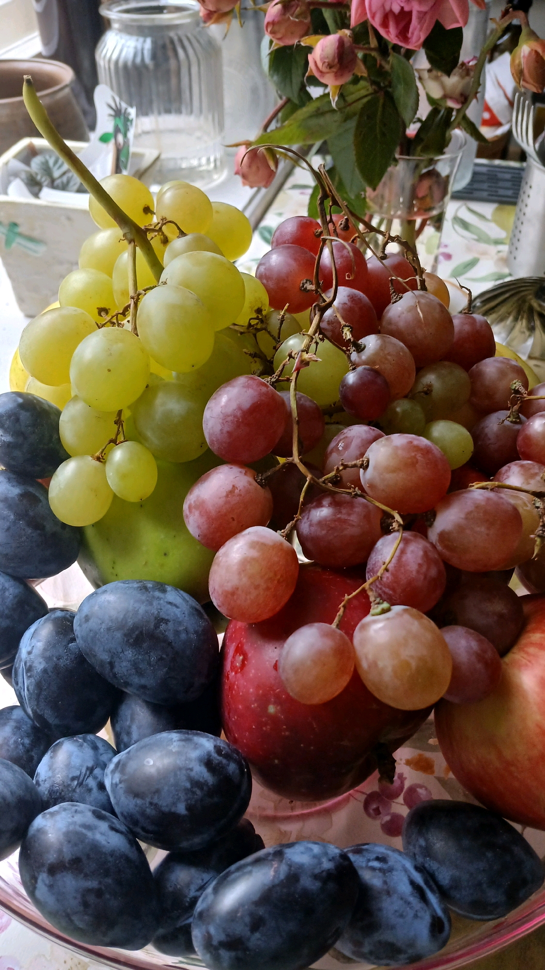 A still life arrangement of fruits, predominantly grapes and plums, is presented in a clear glass bowl. The bowl, filled with a colorful assortment of red and green grapes, alongside deep blue plums and a red apple, rests on a patterned surface. In the background, out-of-focus elements suggest an indoor setting, possibly a kitchen or dining area, with hints of glassware and foliage. The overall lighting is natural and soft, highlighting the textures and natural sheen of the fruits. The scene is devoid of human presence and activity. No specific location in Visaginas, Lithuania can be identified from the visual information provided.