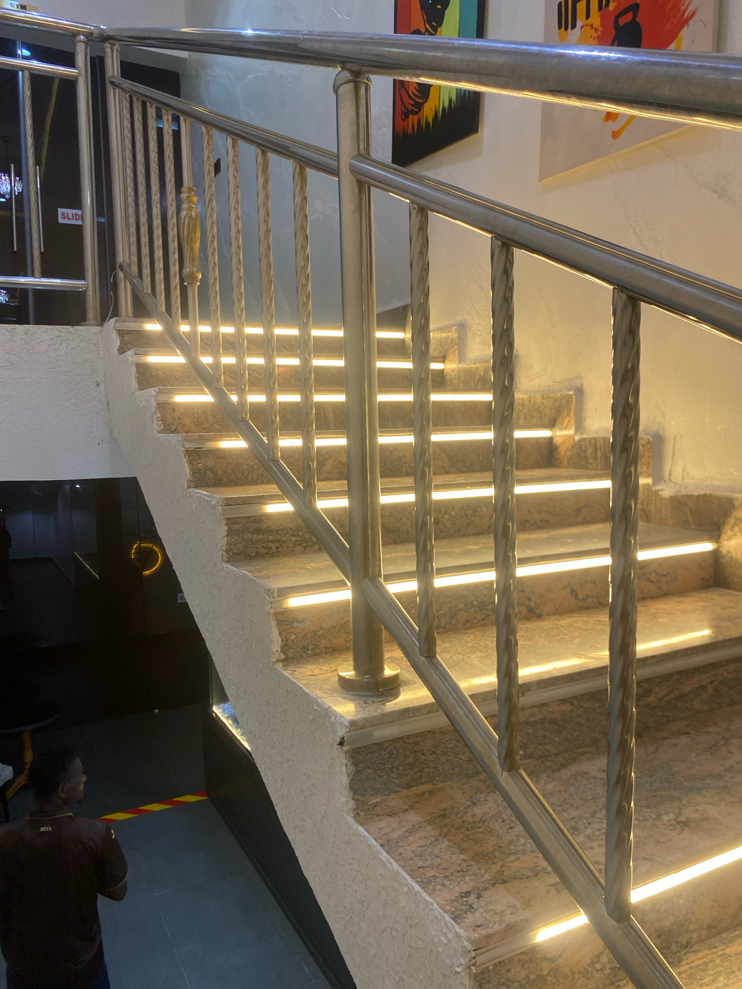A man wearing a maroon shirt with yellow text stands at the bottom of a concrete staircase. The staircase has granite steps and is illuminated by LED strip lights embedded along the front edge of each step. A metal handrail with ornate spindles runs along the side of the stairs. The environment appears to be an indoor space, possibly a gym or a public building, with artwork visible on the wall at the top of the stairs. The city of Barden Yana, Nigeria, is referenced in the prompt but there is no visual information in the image to support this specific location.