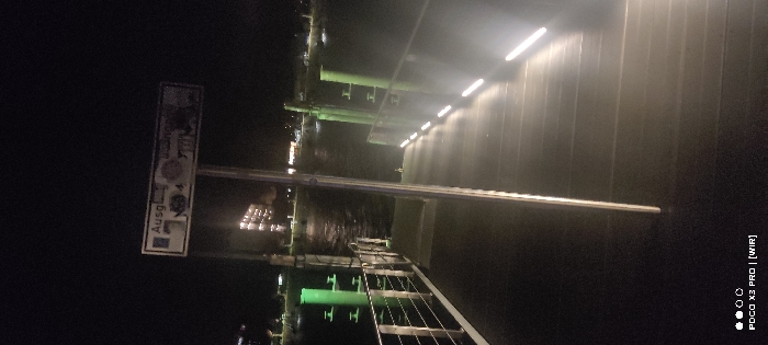 A nighttime image shows a pedestrian bridge with lighting underneath. The bridge's underside is visible, reflecting green light onto the water below. A tall sign on the left displays "Ausgang" with icons. The structure appears modern, with a metal railing and possibly a solid barrier along the walkway. Lights run horizontally above the barrier.