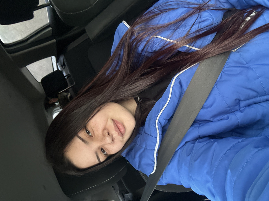 A young adult female is depicted in a close-up, chest-up shot from within a vehicle. She has fair skin, dark brown long hair parted on the left, and visible freckles across her nose and cheeks. Her expression is a slight smile directed towards the viewer. She is wearing a bright blue puffer jacket with white zipper details over a black garment, and a black vehicle seatbelt is positioned diagonally across her chest. The interior of the vehicle shows dark fabric seats, including a headrest behind her and one further back on the passenger side. A blurred figure is partially visible in the rear seat directly behind the front passenger seat. The exterior environment through the vehicle windows is indistinct. The precise location within the city of Padarevo, Bulgaria, cannot be determined from the visual information provided.