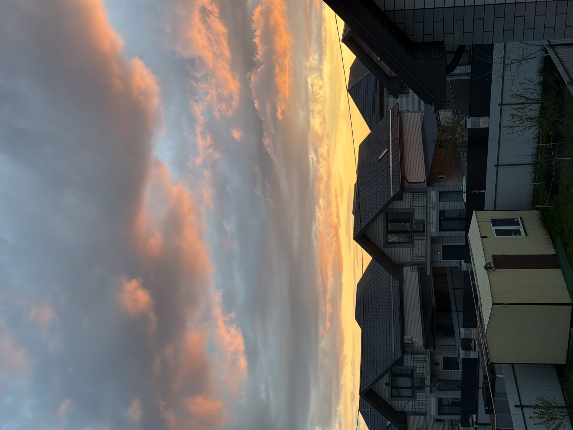 Captured at dusk, the scene presents a low-angle view of a neighborhood in Rossiyskiy, Russia. The sky is dominated by vibrant clouds, exhibiting a gradient of colors from a pale blue-gray to intense oranges and yellows near the horizon, suggesting a sunset or sunrise. A cluster of residential buildings is visible against this dramatic backdrop. The architecture appears modern, with multiple levels and sharply pitched roofs covered in dark tiles. The buildings' facades are a mix of light-colored brick and darker accents, creating a contrast that emphasizes their geometric shapes. The overall environment exudes a sense of tranquility, enhanced by the soft, warm light of the sky and the orderly arrangement of the buildings.