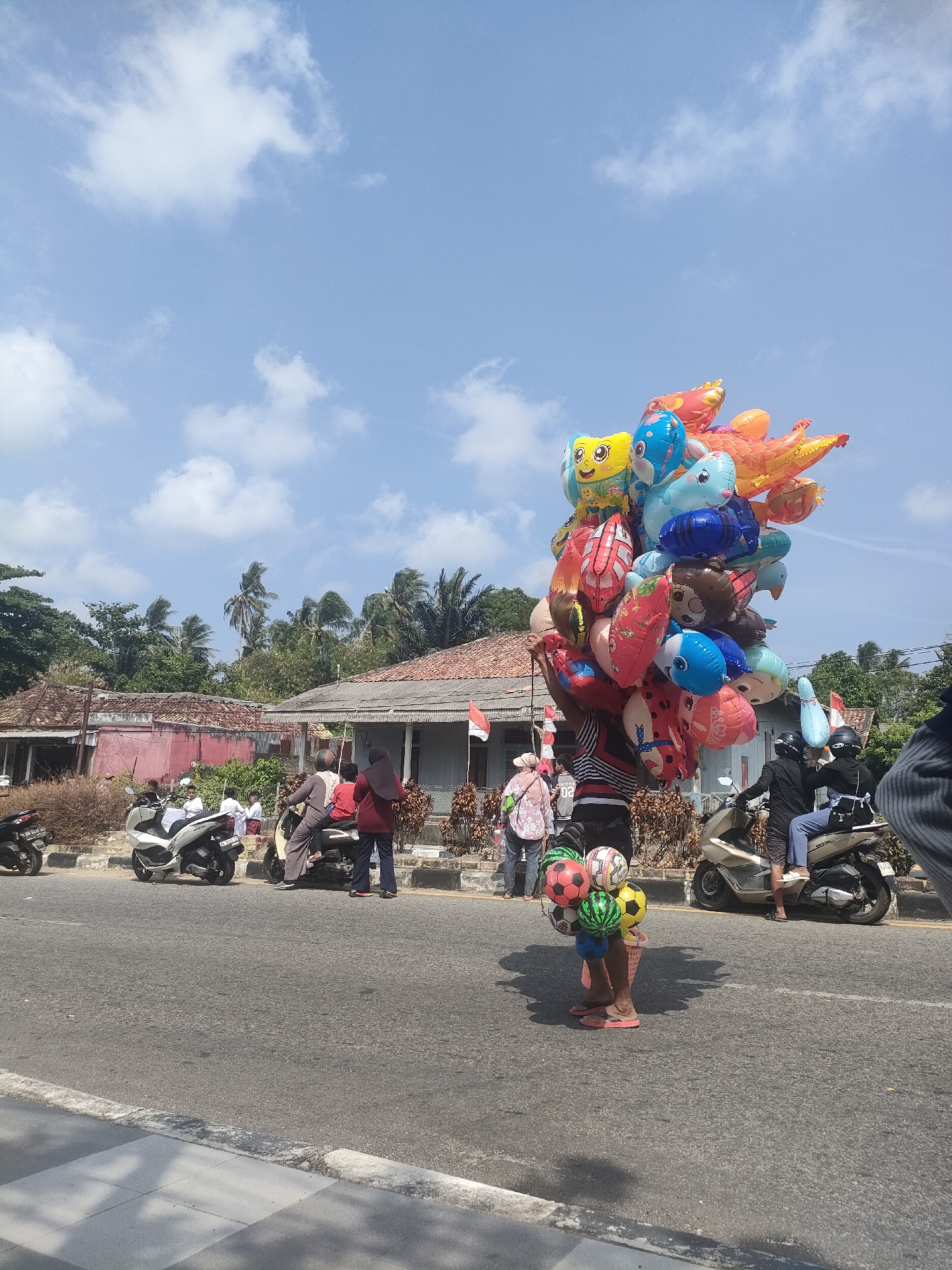 A street vendor stands on the side of a road in Sungailiat, Indonesia, holding a large cluster of brightly colored balloons shaped like animals and cartoon characters. He is also carrying a bucket of soccer balls. The vendor is wearing a striped shirt and pink flip-flops. In the background, there are several houses with tile roofs, palm trees, and a few motorcycles parked along the side of the road. A small group of people are gathered near one of the houses, and two people are riding a motorcycle in the foreground. The sky is clear and blue with a few white clouds.