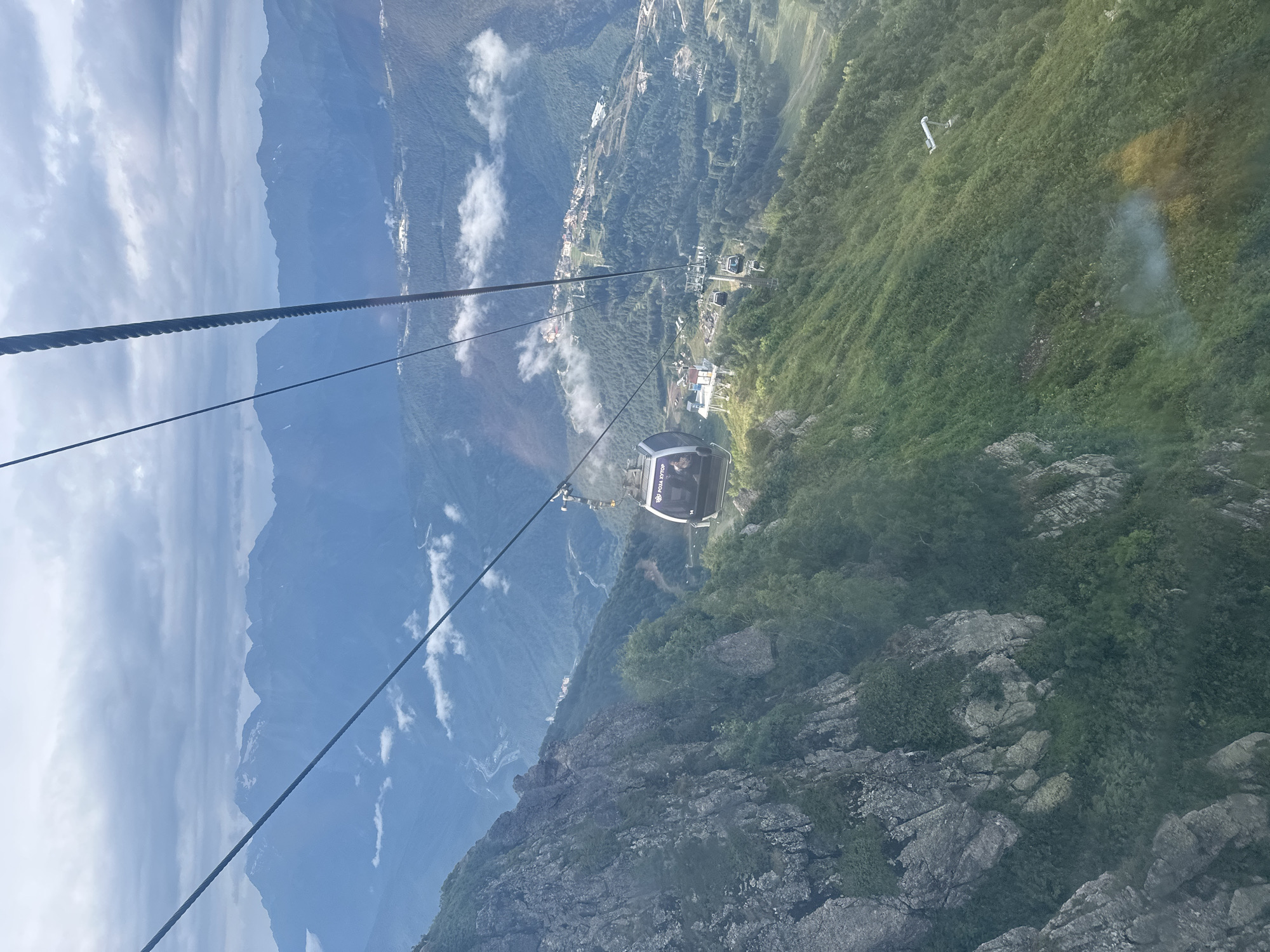 The image captures a view from a cable car ascending a mountain in Estosadok, Russia. The cable car, with the text "РОЗА ХУТОР" visible on its side, is moving upwards against a backdrop of a vast mountain range and a hazy sky. Below, a settlement is spread across the verdant, tree-covered slopes, with roads and buildings dotting the landscape. Rocky cliffs and lush green vegetation dominate the foreground and mid-ground, indicating a mountainous terrain. The atmosphere appears somewhat cloudy, with patches of mist or fog present in the valleys. Several other cable cars can be seen on their respective tracks.