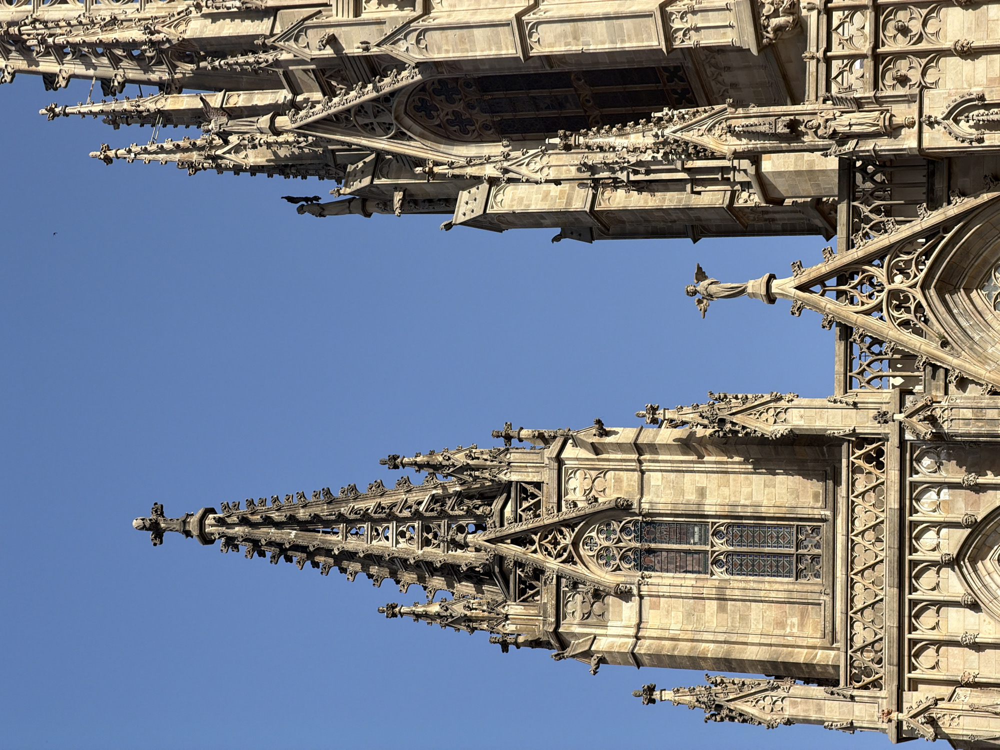 The image shows a close-up, low-angle view of the spires and upper facade of a Gothic cathedral against a clear blue sky. The stone architecture features intricate details, including pointed arches, tracery, gargoyles, and sculpted figures. A small angelic statue stands atop a pinnacle, with wings outstretched. The stonework appears weathered, indicating the age of the structure. The lighting suggests a bright, sunny day. The architectural style strongly suggests this is the Cathedral of Barcelona, located in the Gothic Quarter of the city of Barcelona, Spain.