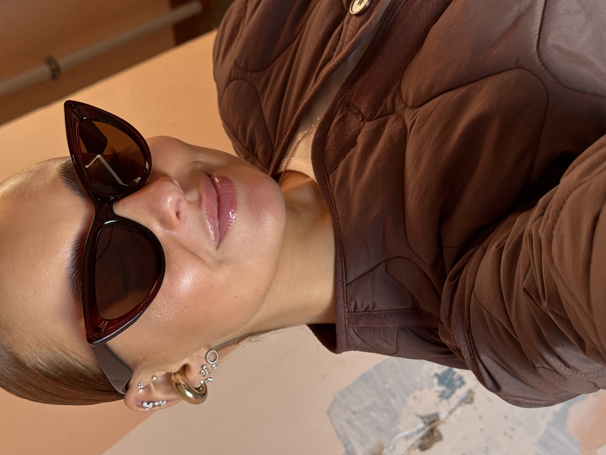 A woman with sunglasses is shown in a close-up, rotated view. She is wearing a brown quilted jacket and has multiple earrings in her left ear. The background appears to be a combination of interior surfaces. There is no verifiable information to support that this image was taken in Chaikovsky, Russia.