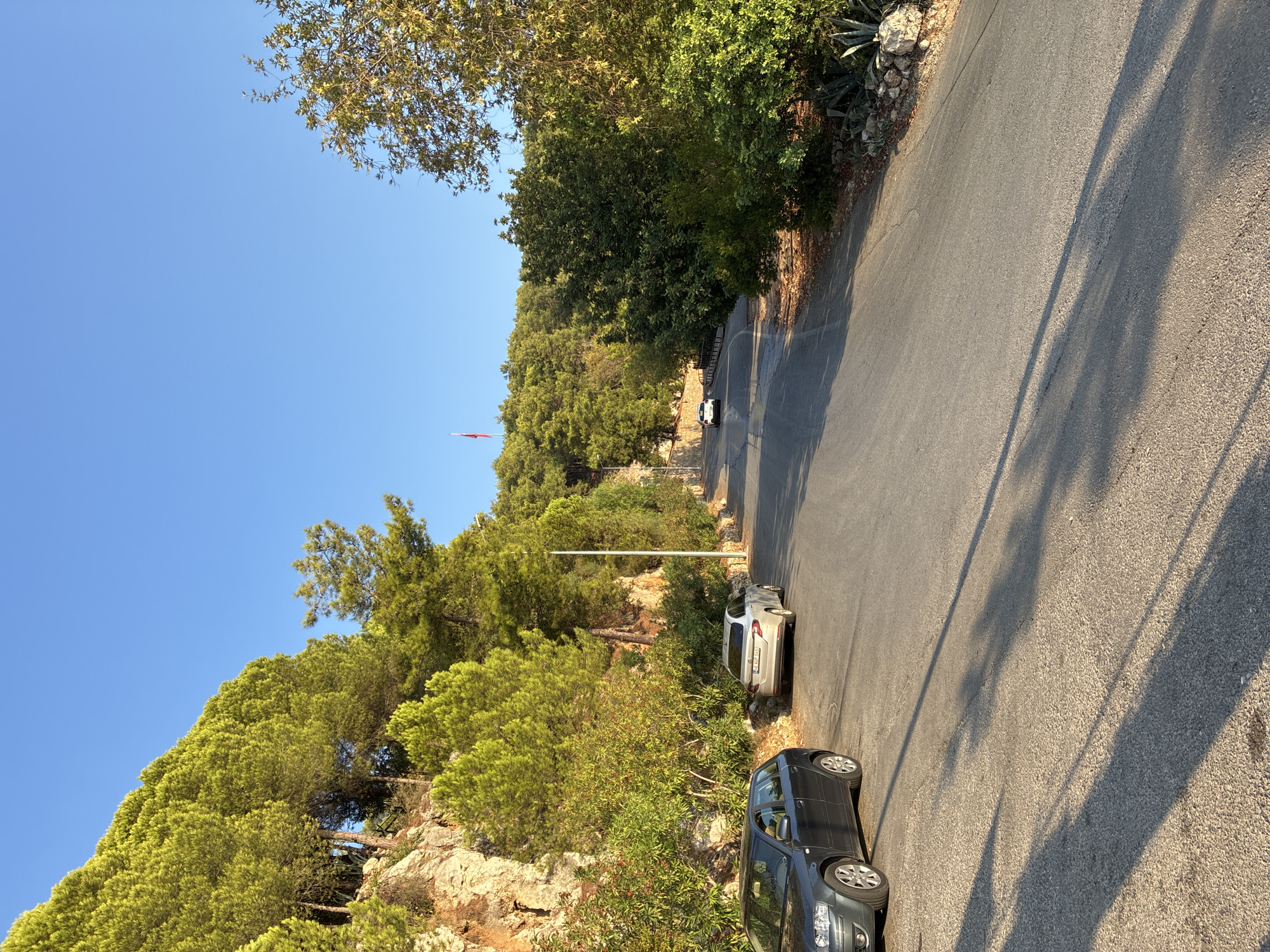 A paved road winds uphill through a forested area with a clear blue sky. On the left side of the road, a dark grey hatchback car is parked on the shoulder. Further up the road, a silver sedan is also parked. A white car is driving up the road in the distance. A flagpole with a red flag is visible on the hilltop. The scene appears to be within or near the city of Alanya, Turkey, possibly on the road leading up to Alanya Castle, given the landscape and the presence of a flagpole. The vegetation consists of pine trees and scrubby bushes. The sun is casting long shadows across the road, indicating it is likely late afternoon or early morning.