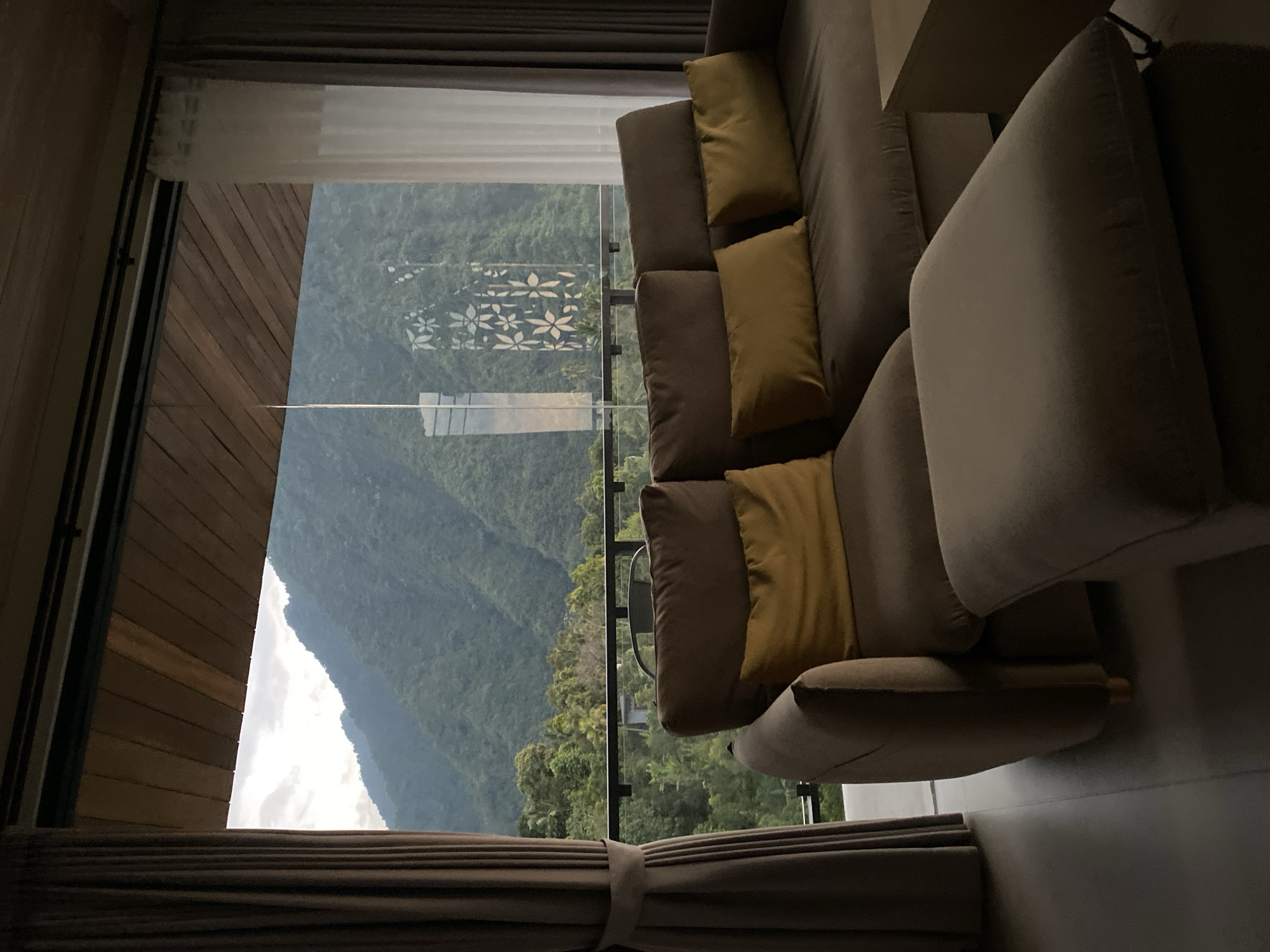 The image depicts an interior view of a room in Sawan, Indonesia, featuring a large, comfortable sofa with several yellow throw pillows. To the left, a brown curtain is partially drawn, and to the right, a sheer white curtain is visible. The room opens up to a breathtaking vista of lush, verdant mountains and dense forest. A railing lines the balcony or terrace, beyond which the natural landscape unfolds. The ceiling of the room is constructed of wooden planks, adding a warm, natural element to the interior design. The overall impression is one of a serene and luxurious retreat, offering a stunning connection to the surrounding natural beauty.