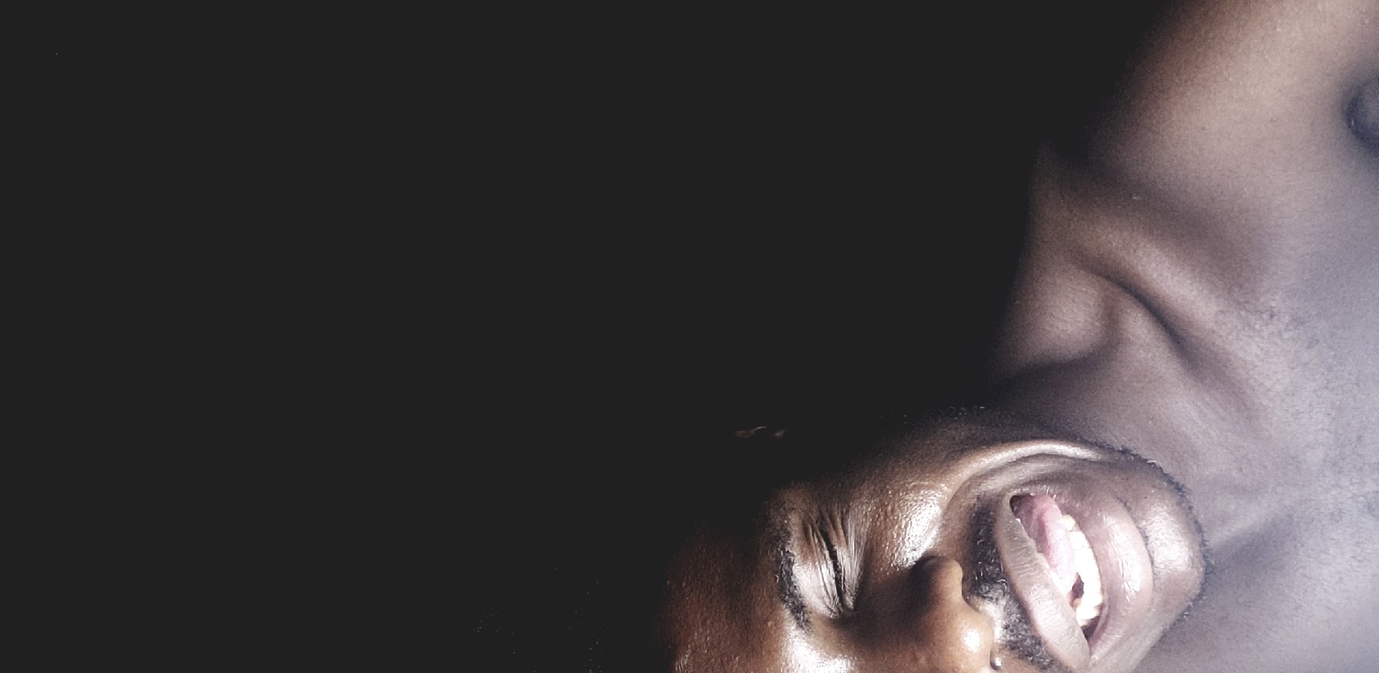 A close-up, low-angle shot shows the face and shoulder of a Black man. The man's skin is dark and appears slightly glossy. His eyes are closed, and his mouth is open, revealing his teeth and tongue. The background is dark and out of focus. There is no indication in the image that confirms its location in Narayi, Nigeria.