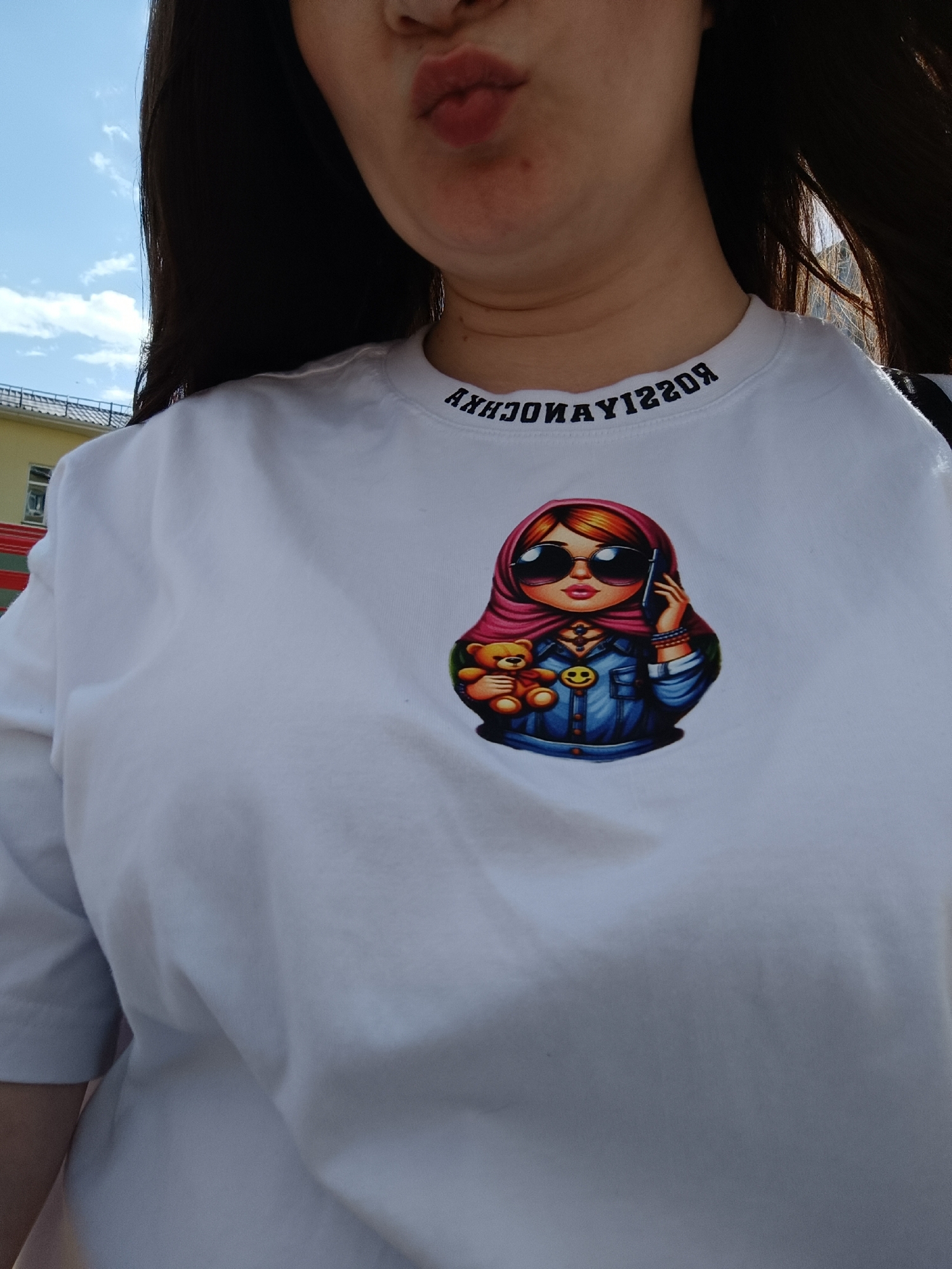 A woman wearing a white t-shirt with a cartoon of a person holding a teddy bear is located in Tobolsk, Russia. The woman is slightly out of focus, but her lips are pursed in a pouting expression. The t-shirt design features a figure in a pink headscarf and sunglasses, holding a teddy bear and a cell phone. The t-shirt also has text around the neckline that reads "ANHCHODNAYT220R" in reverse. A building can be seen blurred in the background. The sky is partially visible in the upper left corner, appearing bright with light clouds.