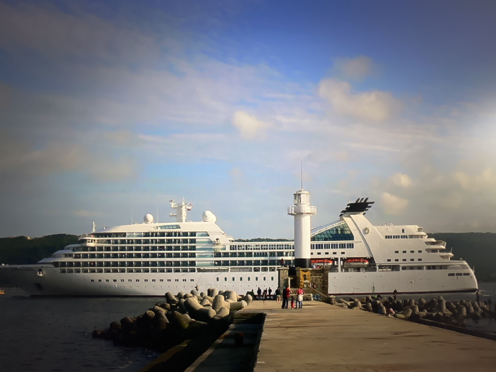 The image captures a cruise ship docked in the port of Varna, Bulgaria. The ship is white with multiple decks and a distinctive modern design, featuring large windows and some green-tinted glass structures. In the foreground, a concrete pier leads towards the ship, lined with tetrapods, which are concrete structures used for coastal defense. A white lighthouse stands prominently on the pier, near the ship. Several people are visible on the pier, some appearing to be passengers or crew, with a few gathered near the lighthouse. The water around the ship is calm, and the sky above is partly cloudy with blue patches. In the background, lush green hills are visible, suggesting the ship is docked near a coastal area. The overall scene depicts a serene moment at a busy port, with the impressive cruise ship as the central subject.
