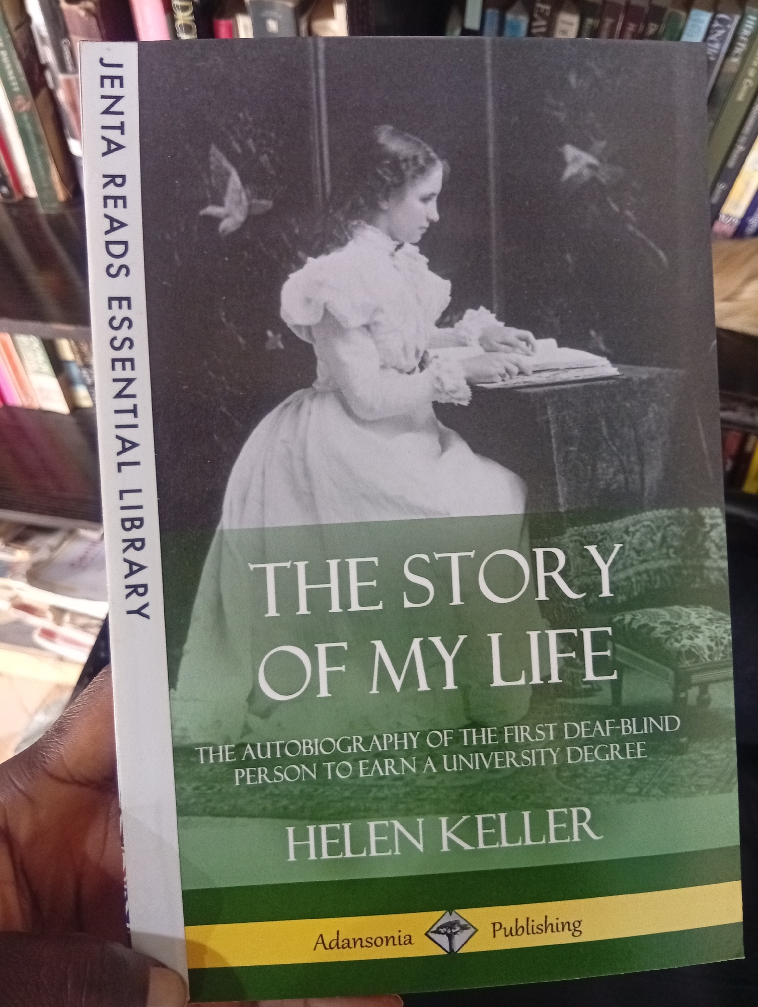 The media file is a photograph of a book cover. The cover features a black and white illustration of a young woman in a white dress sitting at a table with a book. The book title is prominently displayed in large white letters: "THE STORY OF MY LIFE". Below the title, it reads: "THE AUTOBIOGRAPHY OF THE FIRST DEAF-BLIND PERSON TO EARN A UNIVERSITY DEGREE". The author's name, "HELEN KELLER", is printed below this in large letters.
A vertical strip on the left side of the cover has text that reads: "JENTA READS ESSENTIAL LIBRARY". The bottom of the cover has a green band with text "Adansonia Publishing" and a small diamond-shaped logo. The book is held by a person's hand, with dark skin visible. In the background, bookshelves filled with books can be seen, suggesting the photo was taken in a library or bookstore. The overall impression is of an academic or historical subject. The image is shot from a slightly high angle. No specific time of day, weather, or emotional cues are discernible beyond the static nature of the book cover. The location context of Jos, Nigeria is provided separately and not visually evident in the image itself.