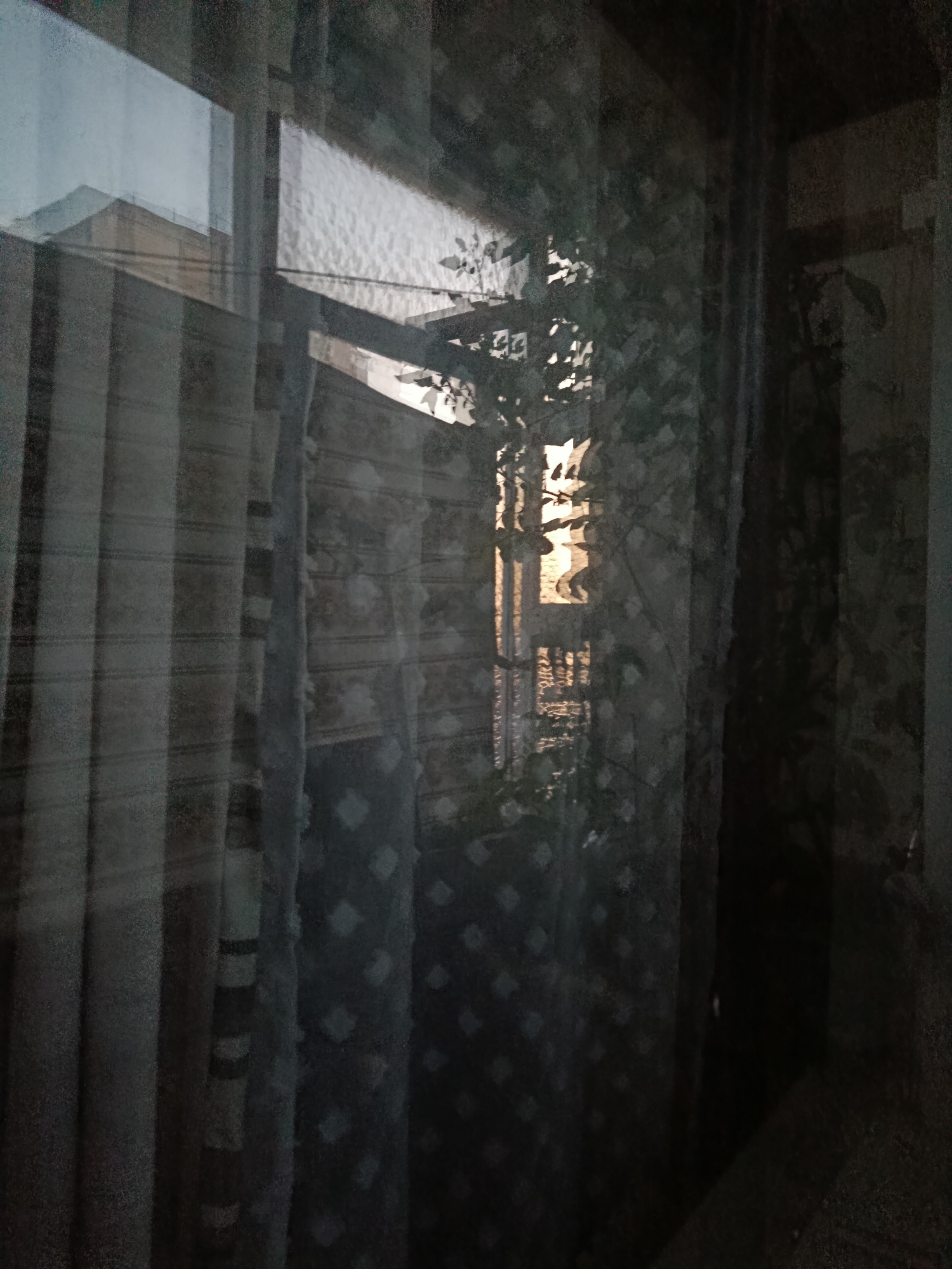 A low-light, slightly out-of-focus image taken indoors, looking out a window. Reflections on the glass obscure the view, showing interior curtains and a hint of outdoor scenery. On the left, vertical striped curtains are visible. To their right, a patterned curtain with a repeating diamond motif is present. The window itself shows a reflection of the sky, a building with a triangular roof, and a tree with sunlight filtering through its leaves. The overall atmosphere is dim and somewhat hazy. The image is tagged as being taken in Cherkaske, Ukraine, suggesting the view is from a window in that location. No people or specific actions are discernible.