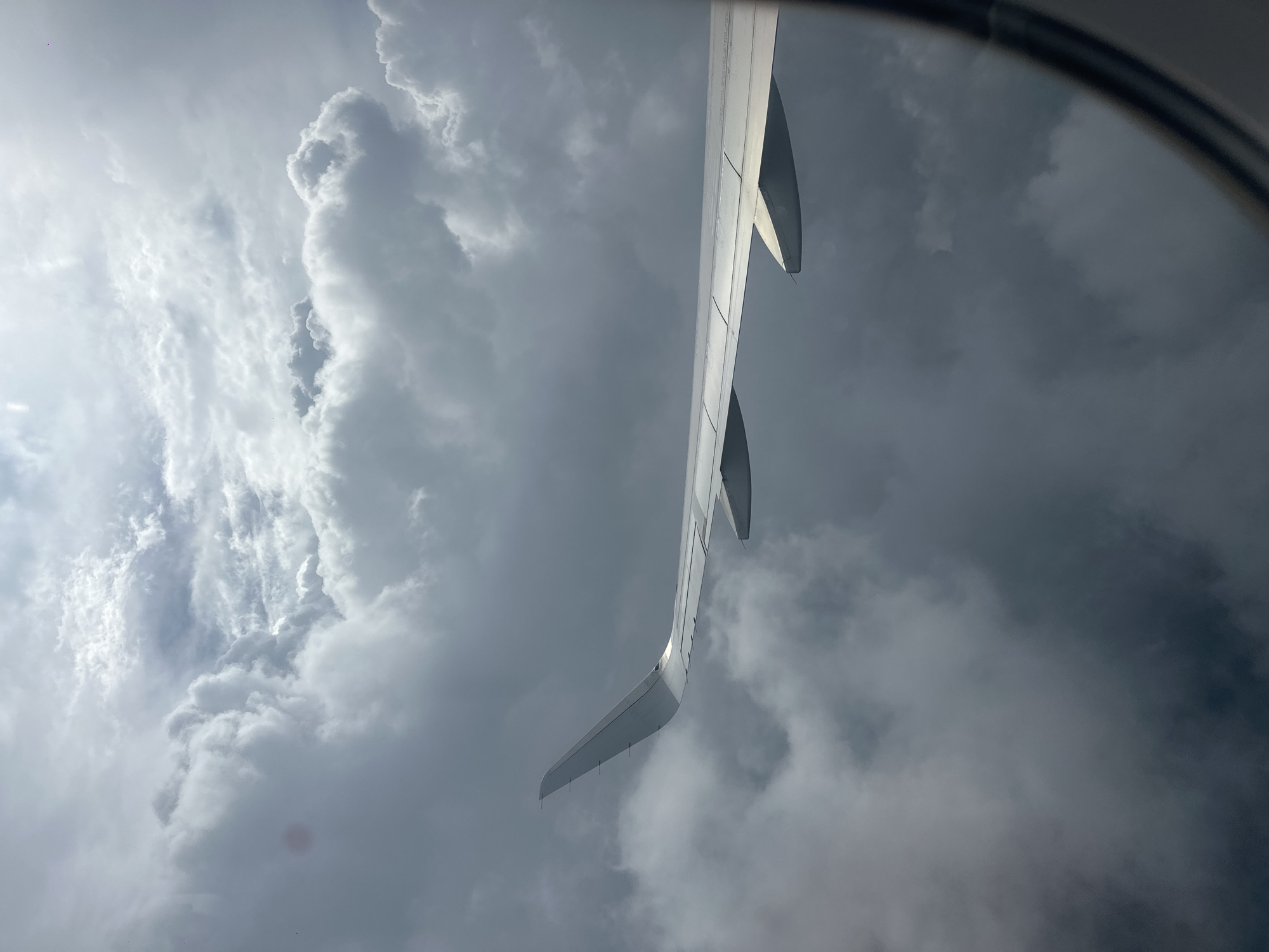The image shows an airplane wing and turbulent clouds from a window. The scene is from an airplane in flight over Fogang County, China. The clouds are gray and dense, with some patches of lighter sky visible. The airplane wing is white and extends across the frame. The image is taken from inside the airplane, looking out the window.