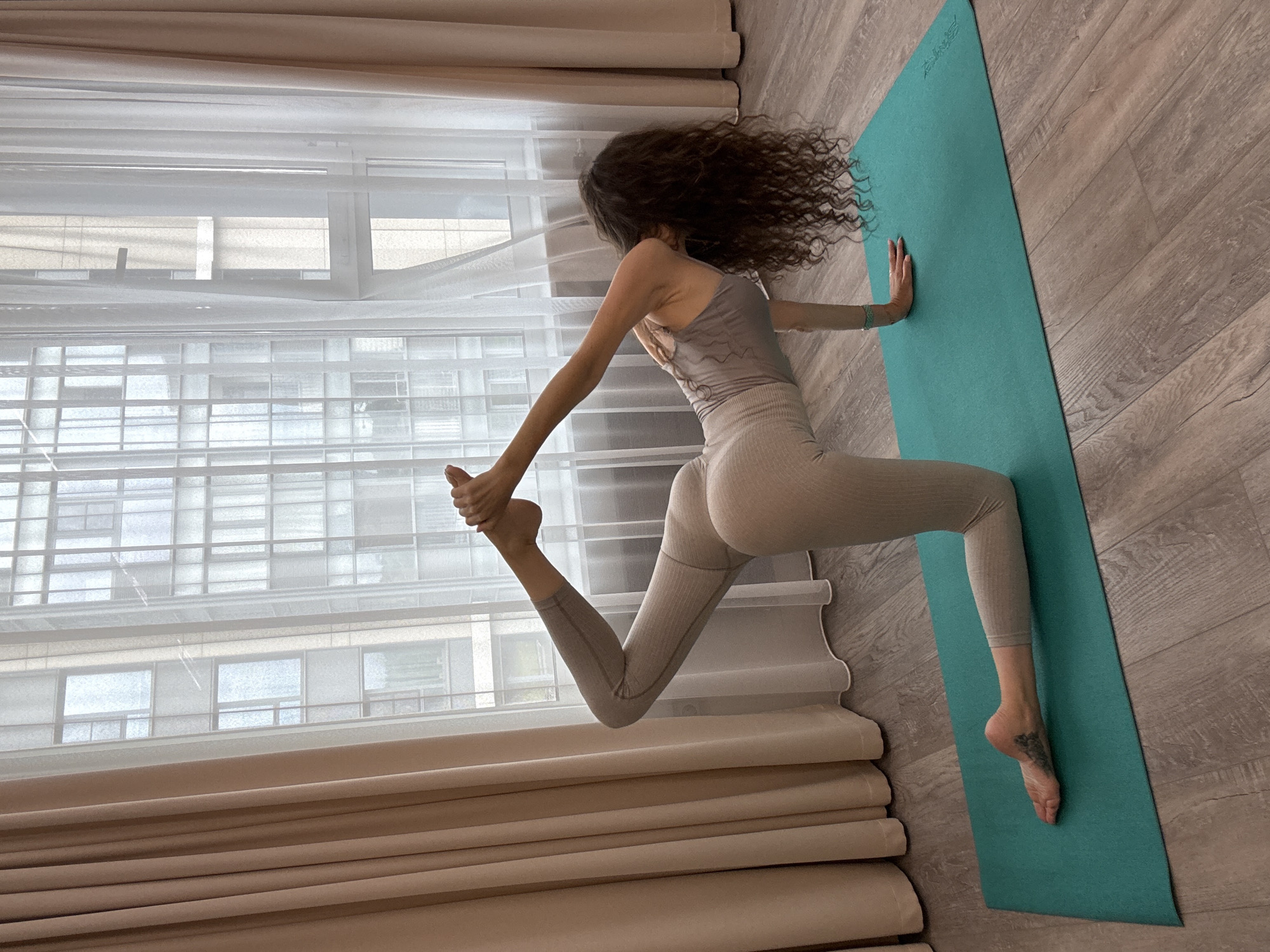 A person is practicing yoga in a room with a large window. The person, with long, curly dark hair, is wearing a light-colored yoga outfit and is positioned on a turquoise yoga mat. They are performing a pose where their right leg is extended forward and their left leg is bent, with their left foot being held by their left hand. Their right hand is resting on the yoga mat for support, and their body is angled towards the window.
The room has light-colored curtains and blinds covering the lower part of the window, through which a building with multiple windows can be seen. The floor is made of light-colored wooden planks. The lighting suggests it is daytime. The overall scene appears to be a personal space, possibly a home, being used for a yoga practice. There is no visible text in the image. The activity is focused and appears to be a moment of physical exertion and concentration.
