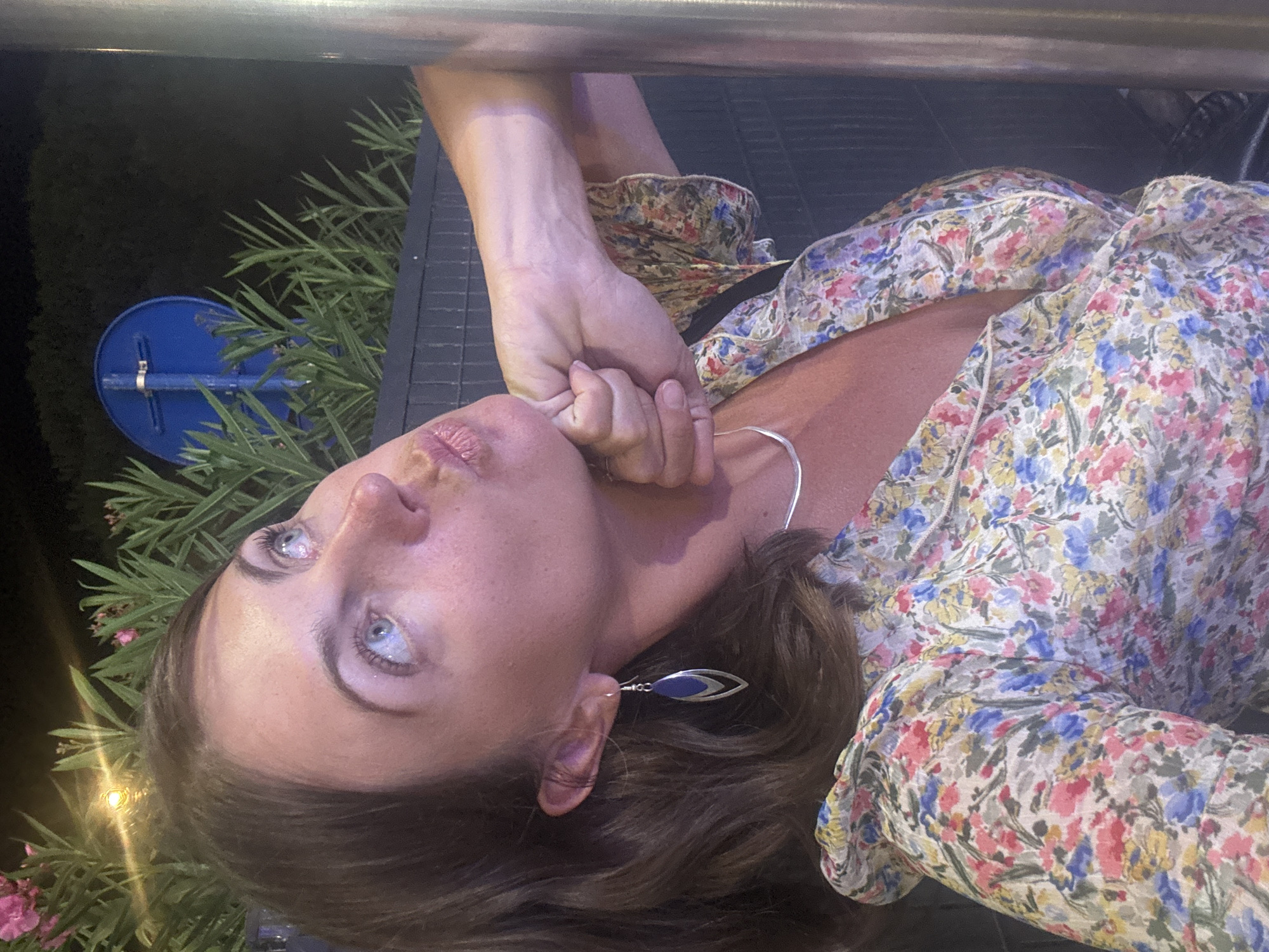 A woman with brown hair and light blue eyes is taking a selfie outdoors at night. She is wearing a floral print dress and a silver necklace. Her left hand is resting on her chin, and she is puckering her lips. Behind her, there is a dark fence and some greenery. To her left, a blue circular sign is visible. The scene appears to be in Argelès-sur-Mer, France, given the mention of the location.