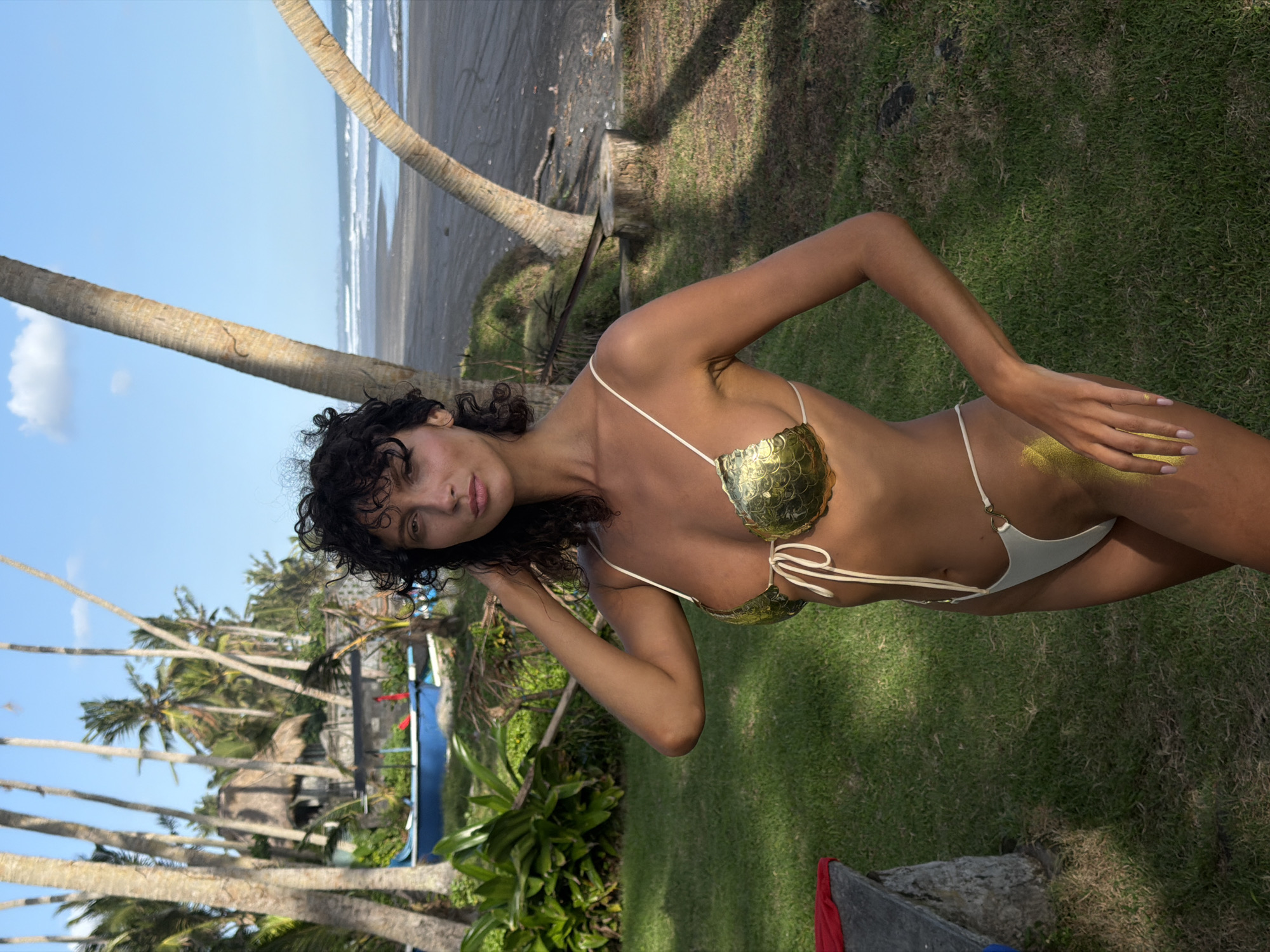 The image is of a woman in a gold-and-white bikini standing on a grassy area near a beach in what appears to be Kerambitan, Indonesia. The woman has curly black hair and is wearing a bikini that has gold-colored, scale-like cups and a white bottom with string ties. She has some gold glitter or powder on her leg and hand.
In the background, there are palm trees, the ocean, and dark sand. Other trees and some blue structures are visible behind her and to the left. The sky is blue and there are signs of a building between trees. The location appears to be a tropical or beach-side environment.