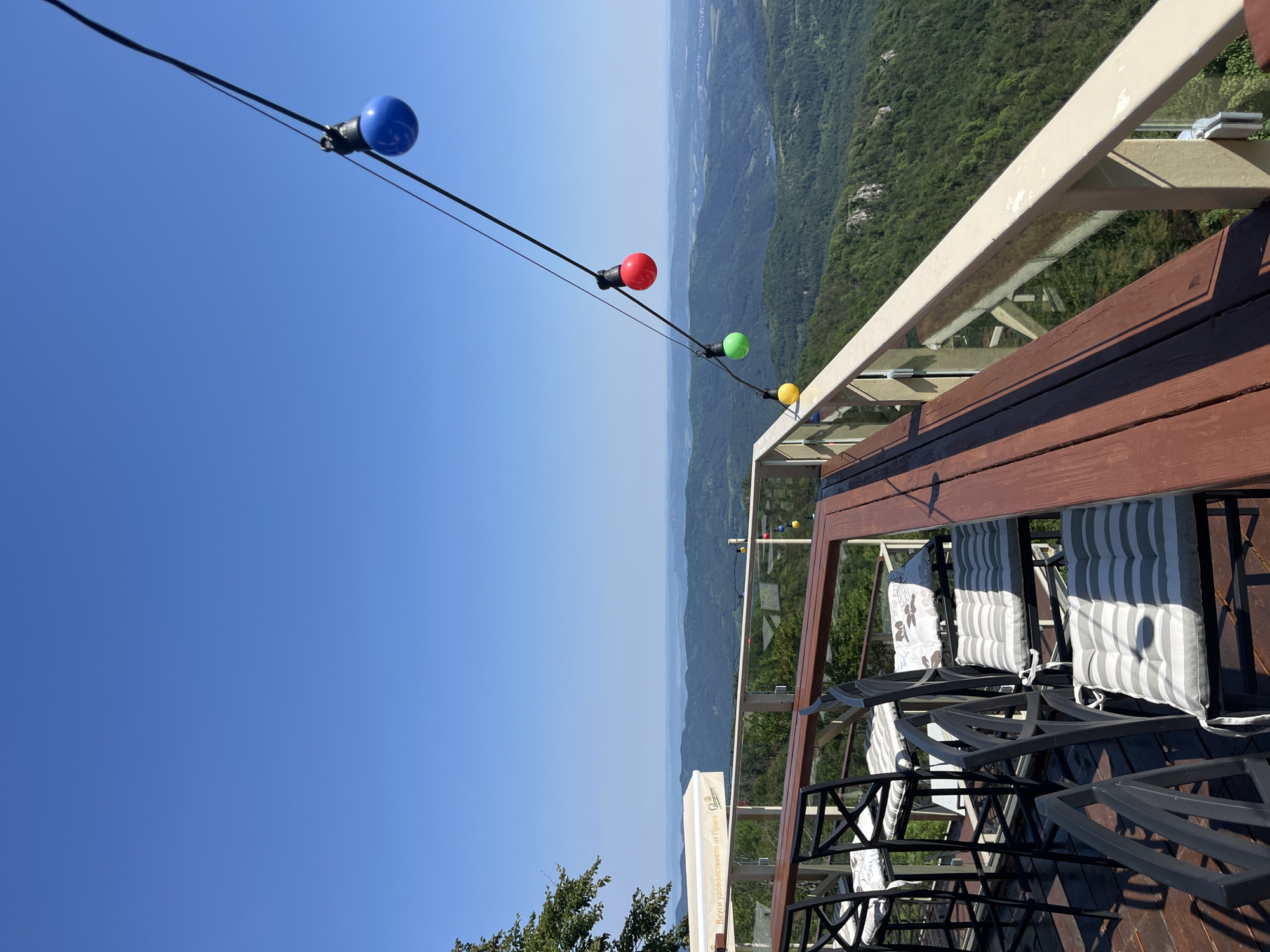 An outdoor seating area on a balcony overlooks a lush, mountainous landscape under a clear blue sky. The balcony has a wooden floor and a light-colored railing with glass panels. Several bar stools with striped cushions are positioned around a wooden table. A string of colorful light bulbs – blue, red, green, and yellow – hangs overhead, attached to a dark cable. In the background, layers of green, tree-covered mountains recede into the distance, with a hint of haze obscuring the farthest peaks. The scene is bathed in bright sunlight, casting shadows on the balcony.